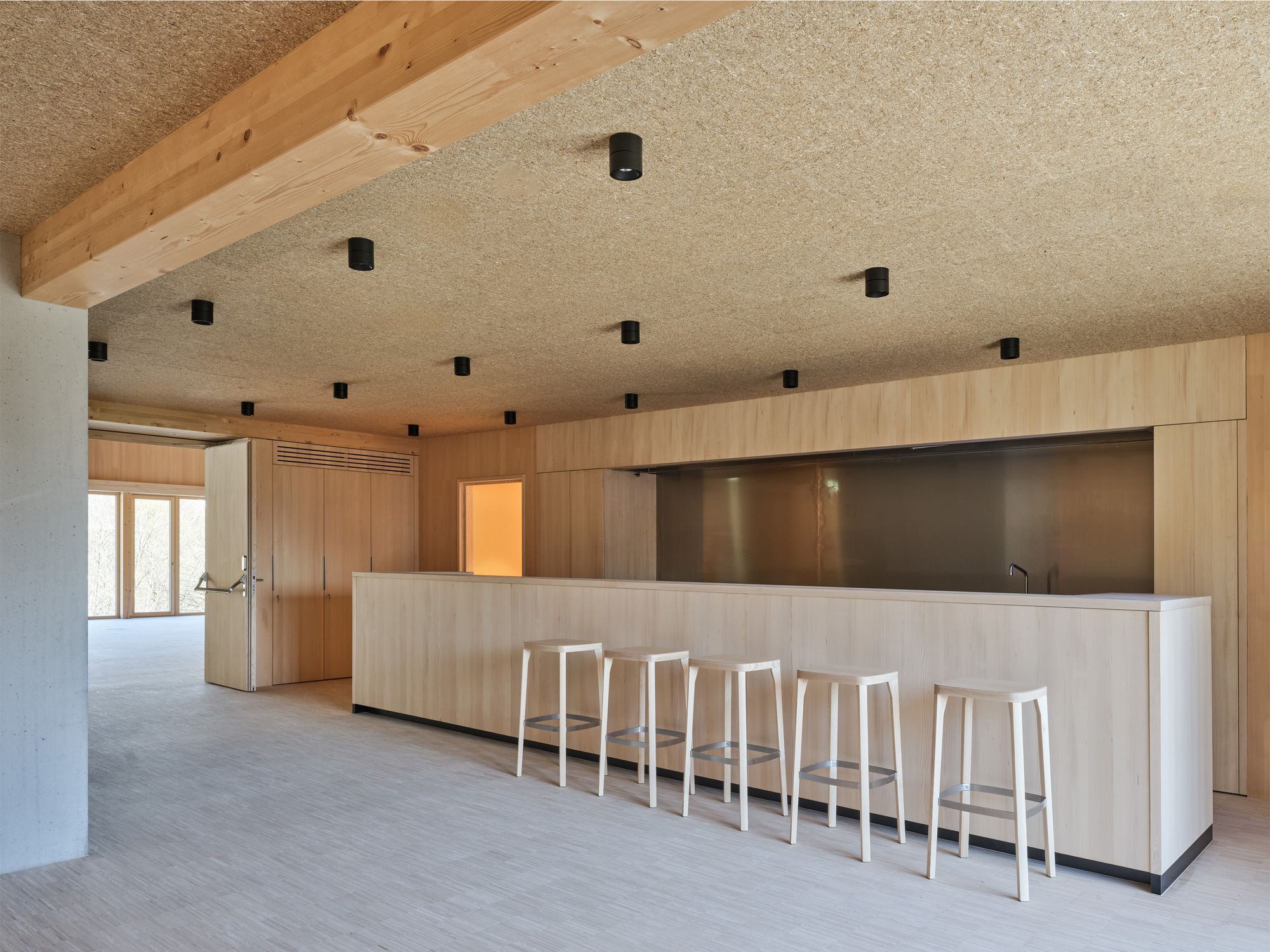 Wood-panelled counter with five pale stools and a steel back panel beneath a ceiling of wood-wool boards and a glulam beam