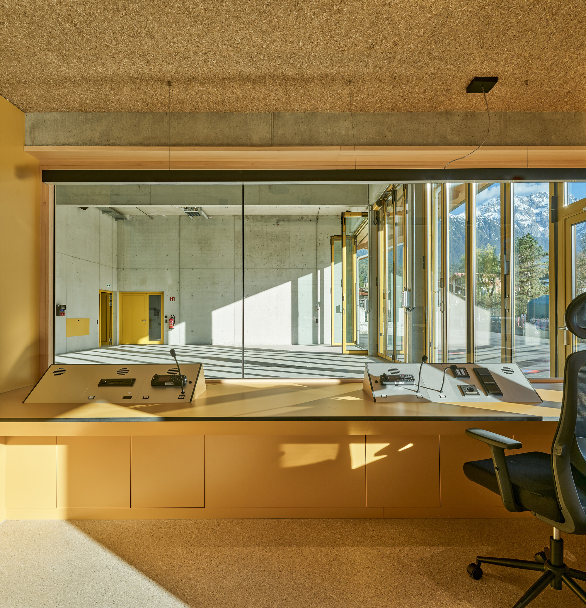 Control desk with microphones behind a glass window overlooking a concrete hall, with sunlit snowy peaks visible through the yellow-framed doors