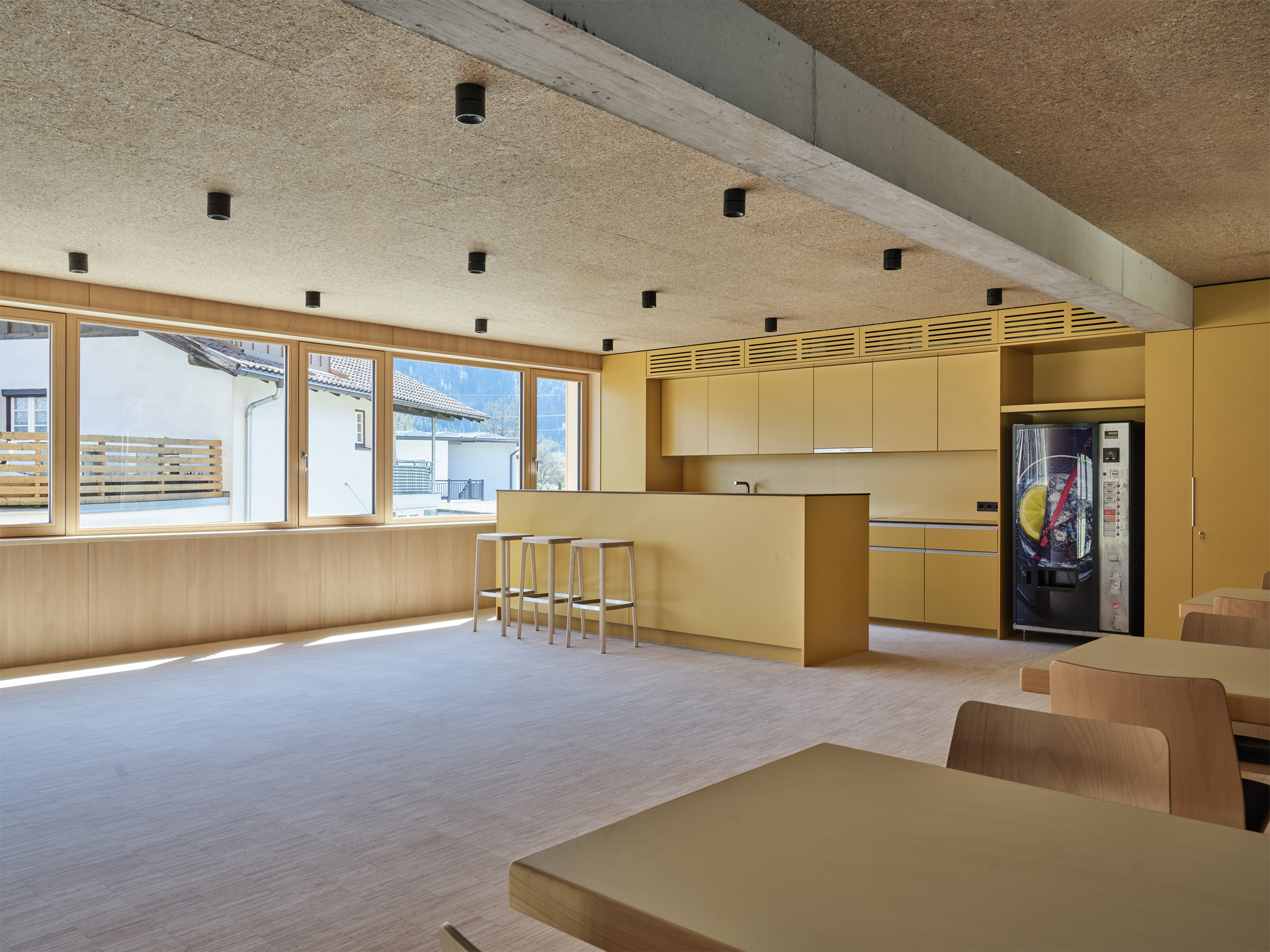 Break room with yellow cabinetry, a freestanding kitchen island, stools, a vending machine and tables, lit by a long window of afternoon light