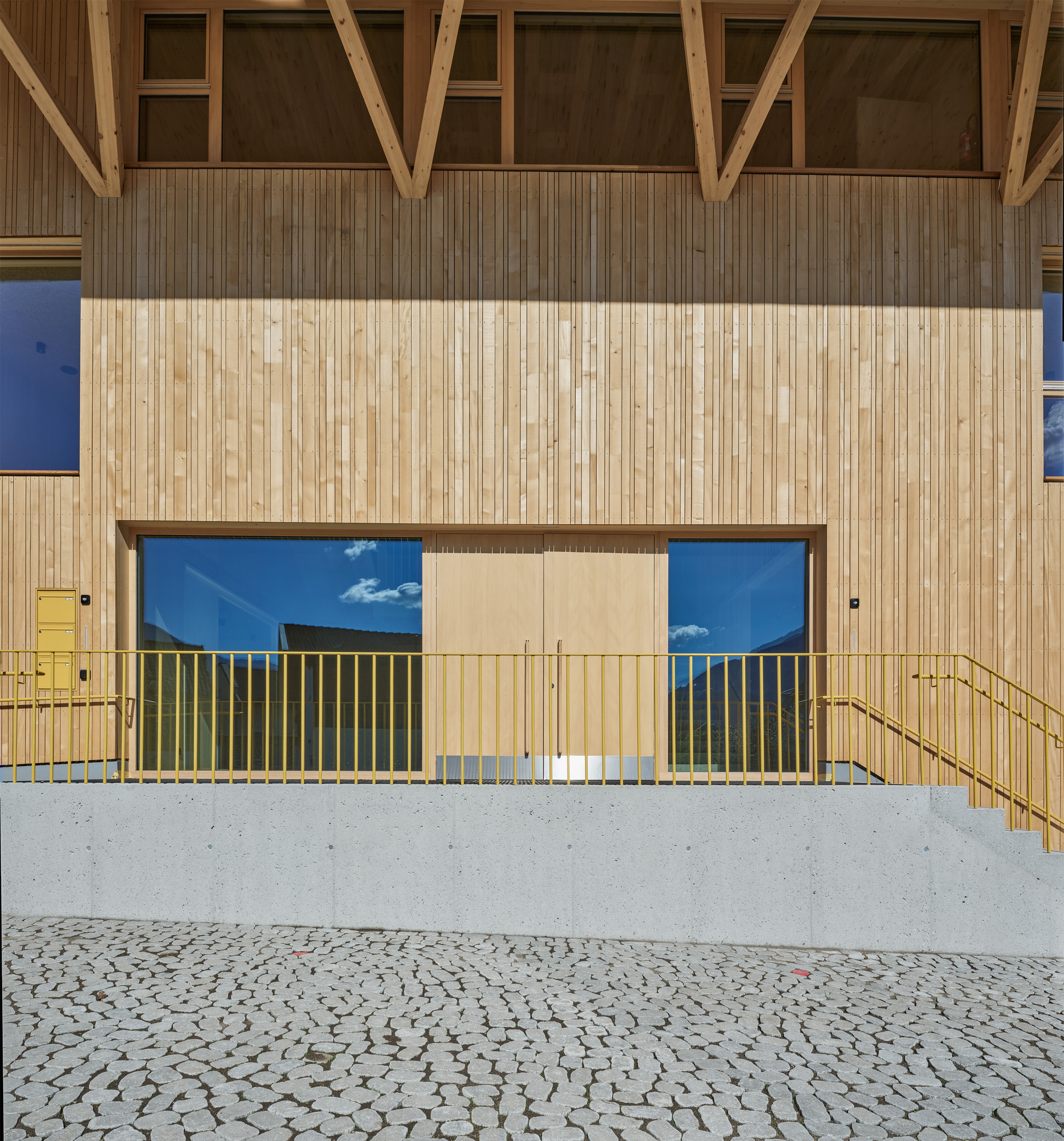 Entrance on the vertical timber facade with a yellow railing and stair above a concrete wall and cobbled paving, sky reflected in the glass