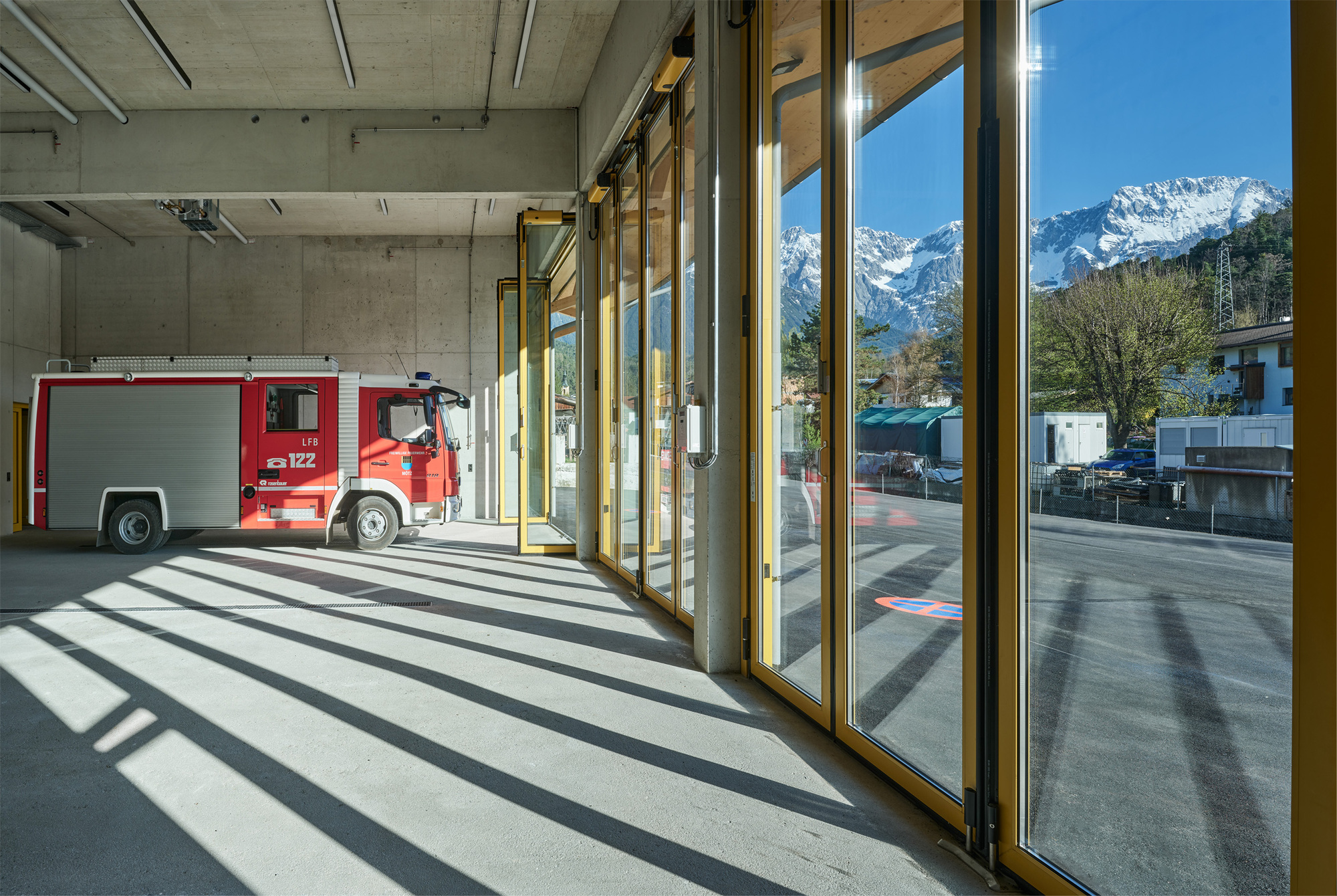 Fire engine parked in a concrete bay behind tall glazed folding doors, with sunlit shadow stripes on the floor and snowy peaks beyond