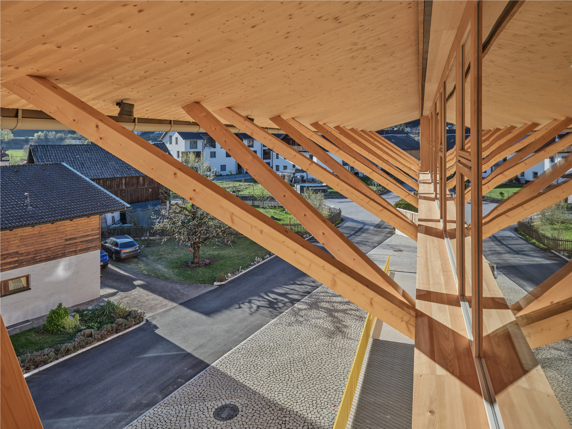 Looking down from under a timber canopy with diagonal glulam struts toward a cobbled forecourt and village street in afternoon sun