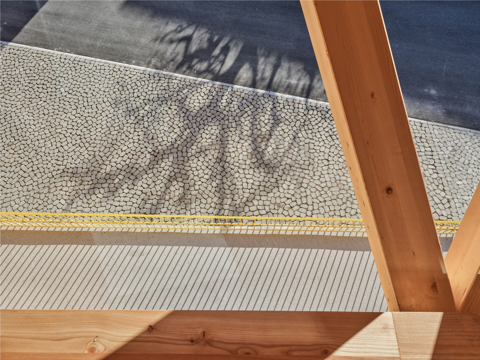 Close-up of mosaic paving and a yellow edge strip seen between timber beams, with tree shadows cast across the stones
