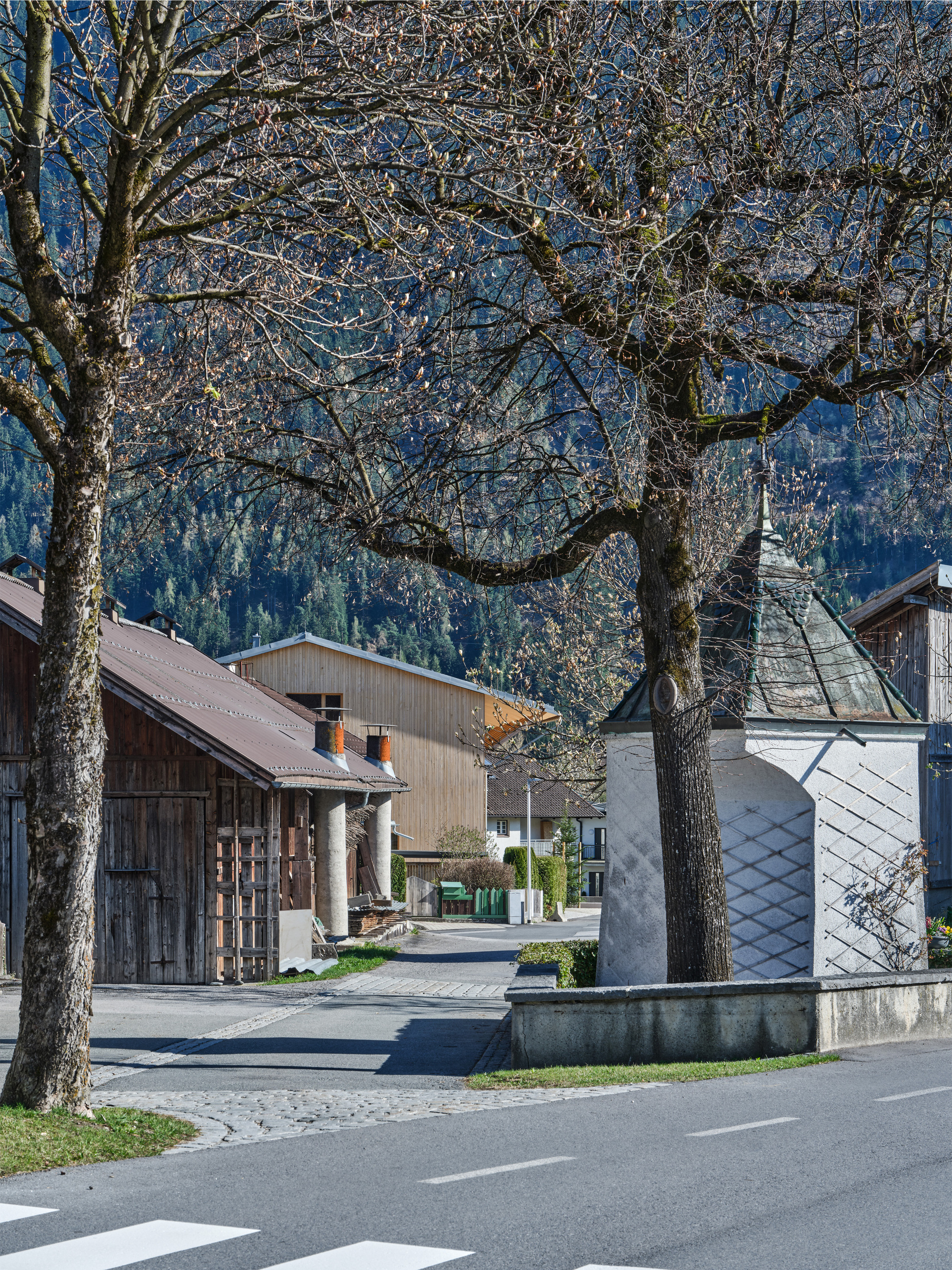 Village street corner with a whitewashed wayside shrine, bare trees and timber barns, a wood-clad building and forested slope behind