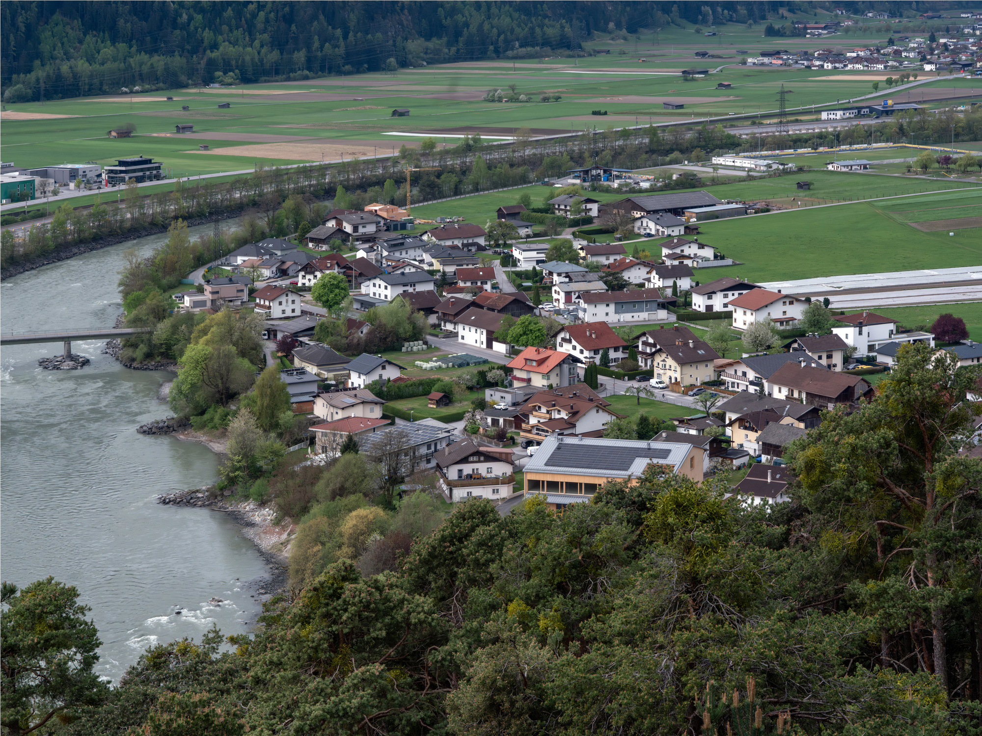 Aerial view of a valley village of pitched-roof houses beside a river, with green fields and conifer-covered slopes in the foreground