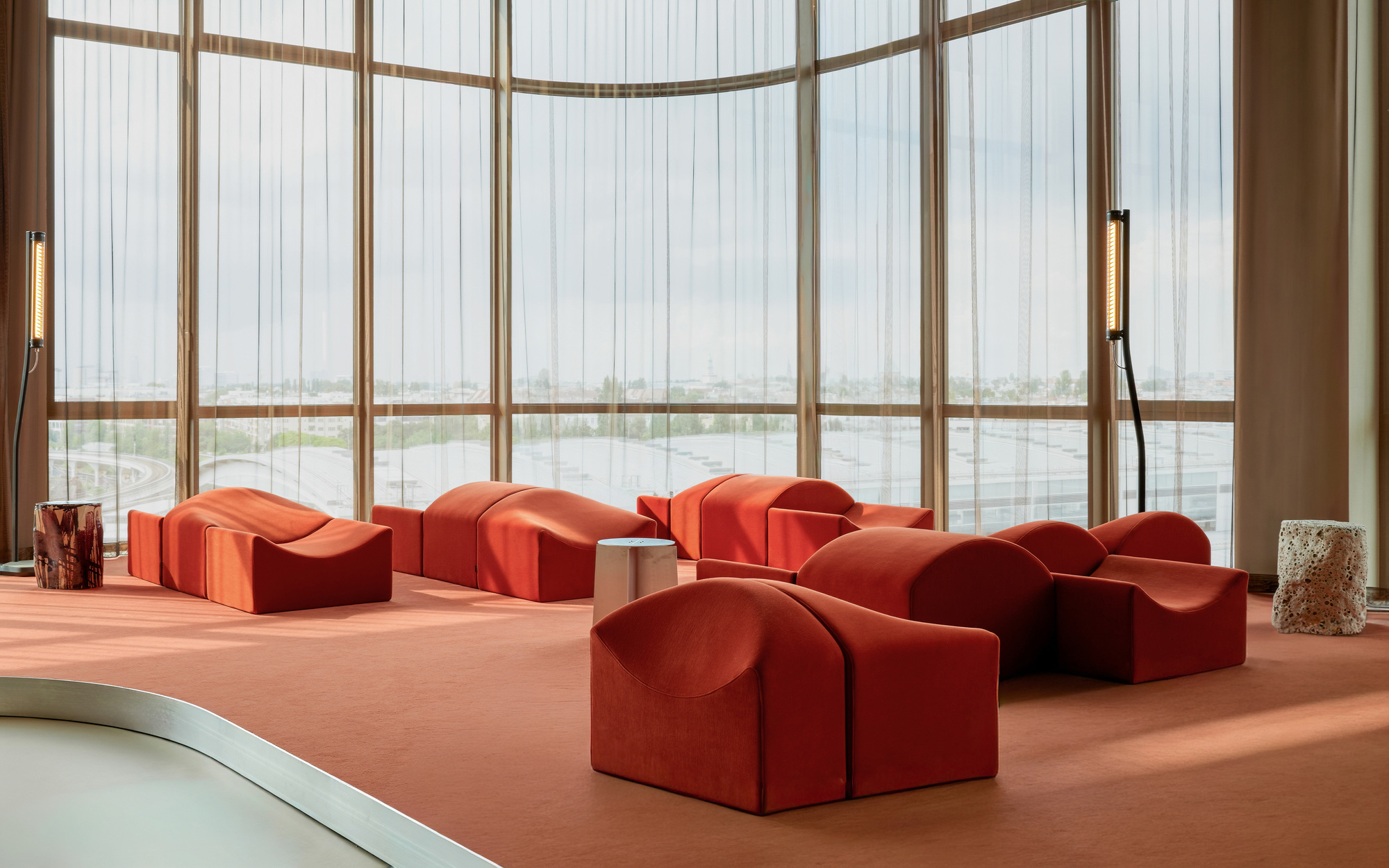 Rust-red sculptural lounge seating on matching carpet before a curved curtain wall of sheer drapes and hazy sky