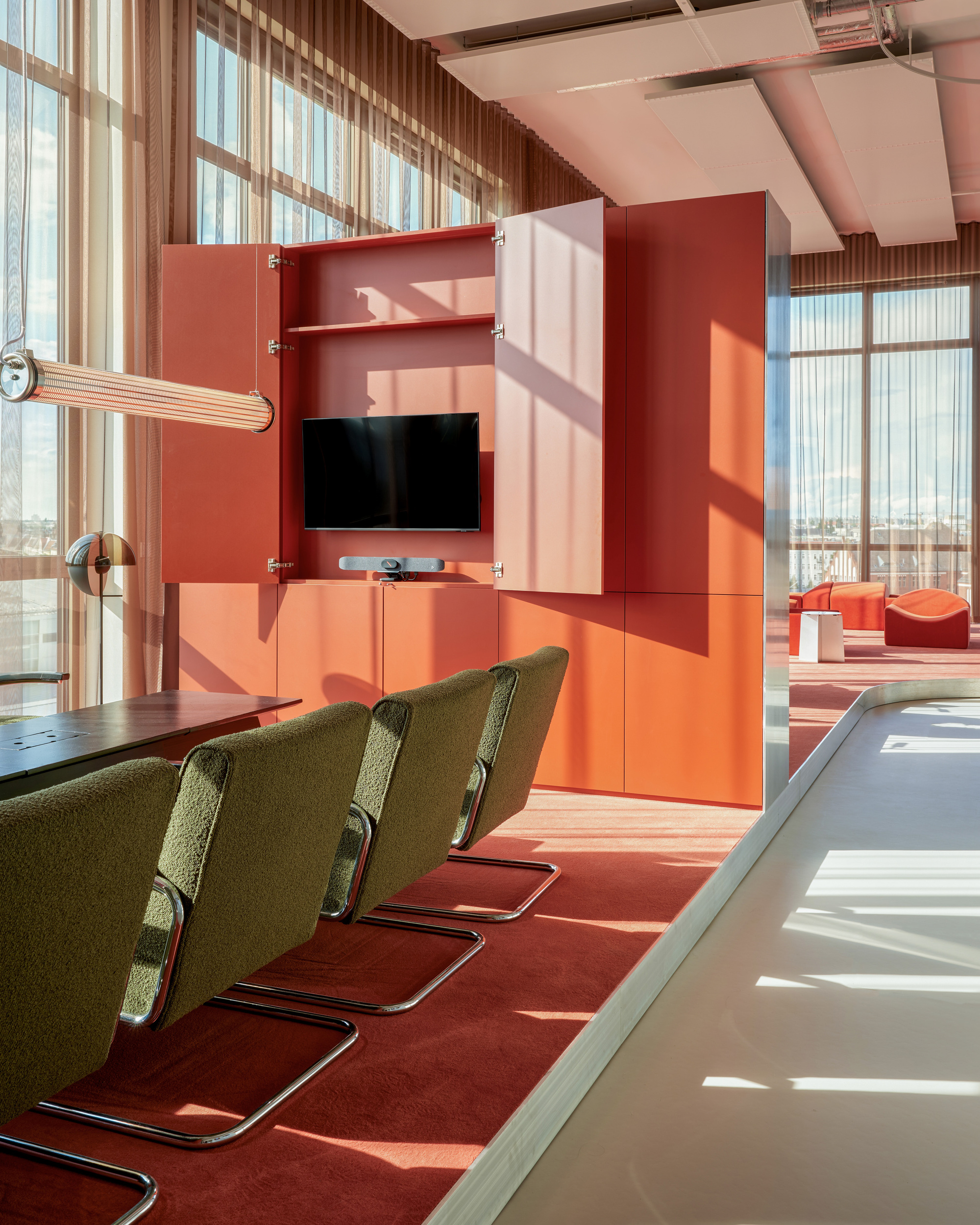 Olive boucle cantilever chairs facing an orange cabinet with open doors and a wall-mounted screen in low sunlight