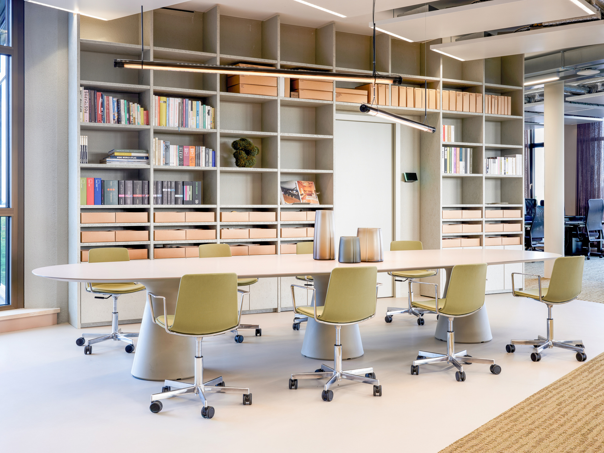 Long white table with olive swivel chairs in front of floor-to-ceiling shelves holding books and cardboard archive boxes