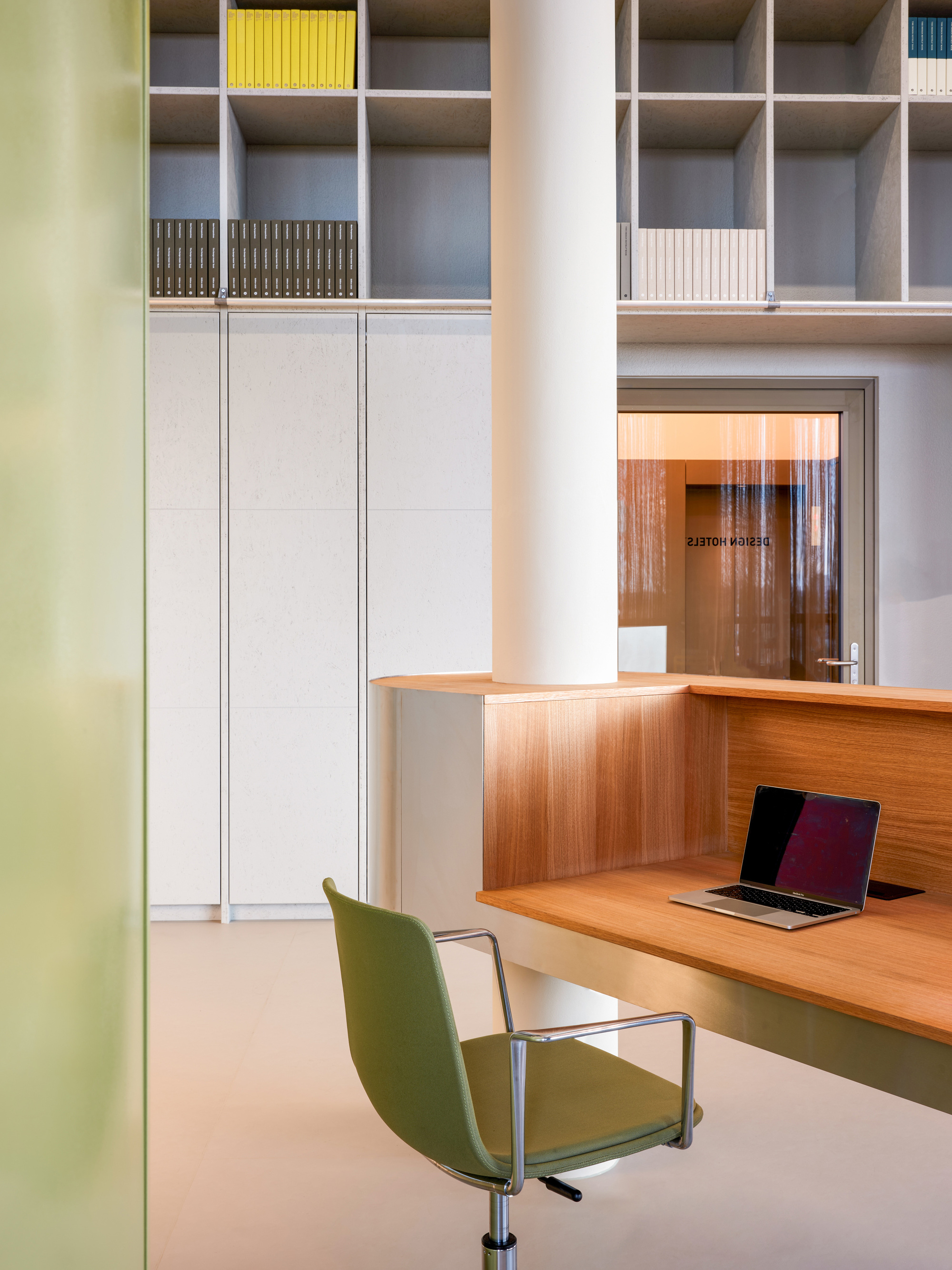 Oak-veneered reception desk with a laptop and green chair beside a round white column and tall shelving