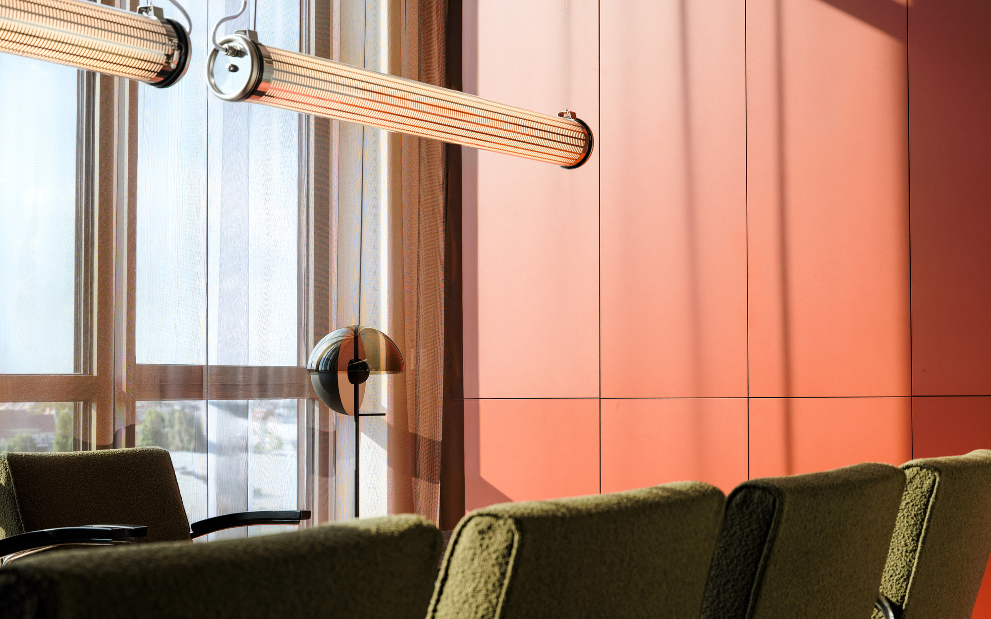 Close view of ribbed tube pendant lights over olive boucle chair backs against an orange panelled wall and sunlit window