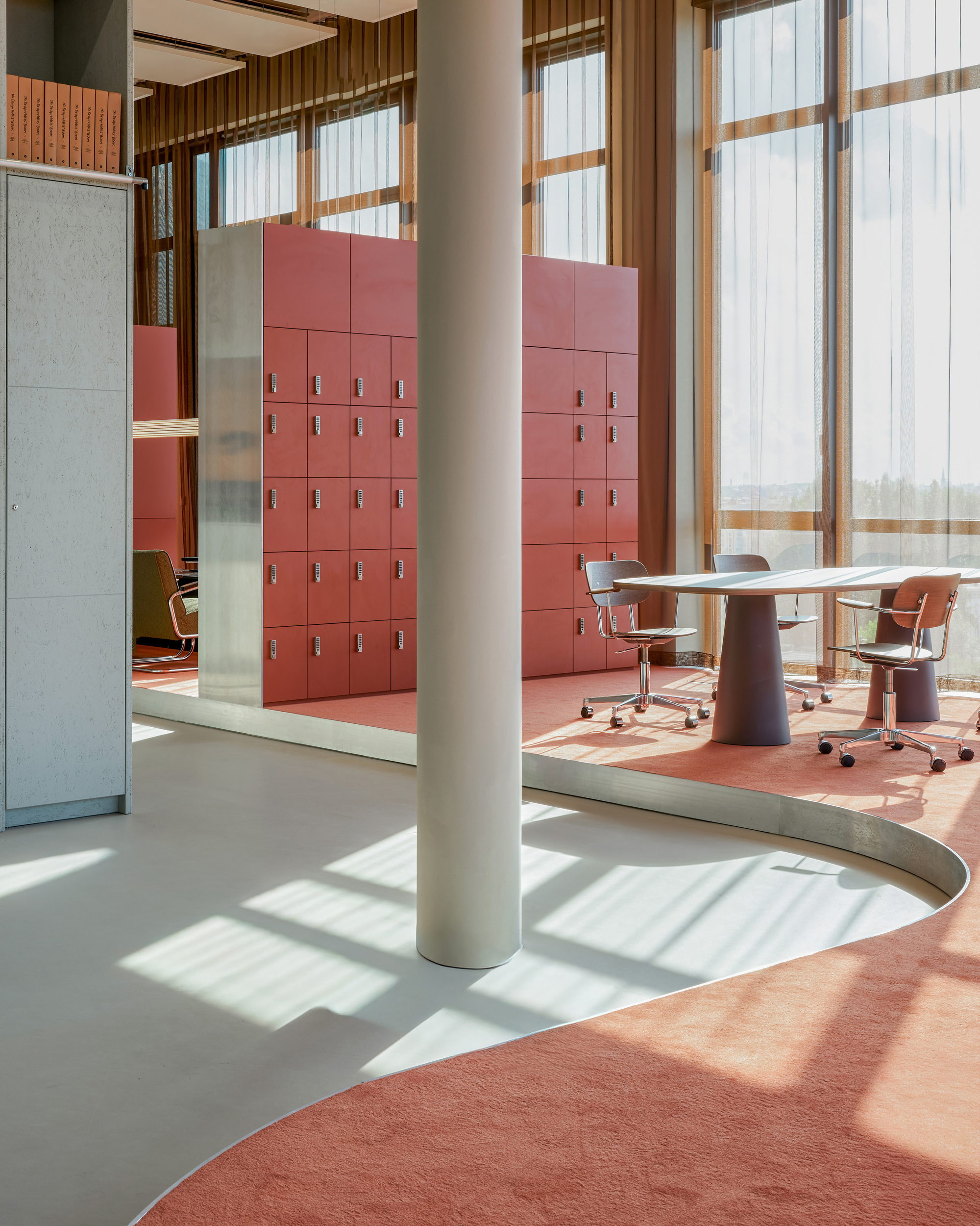 Curved edge of the floor between grey linoleum and red carpet, with a round column, red lockers and a conical-base table in sunlight