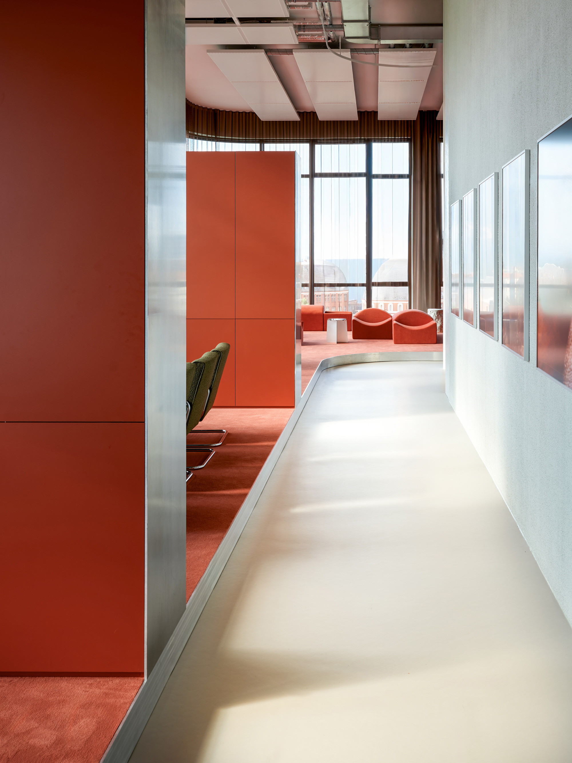 Curving pale corridor running beside red cabinet volumes and a framed wall, leading to red lounge chairs at a tall window