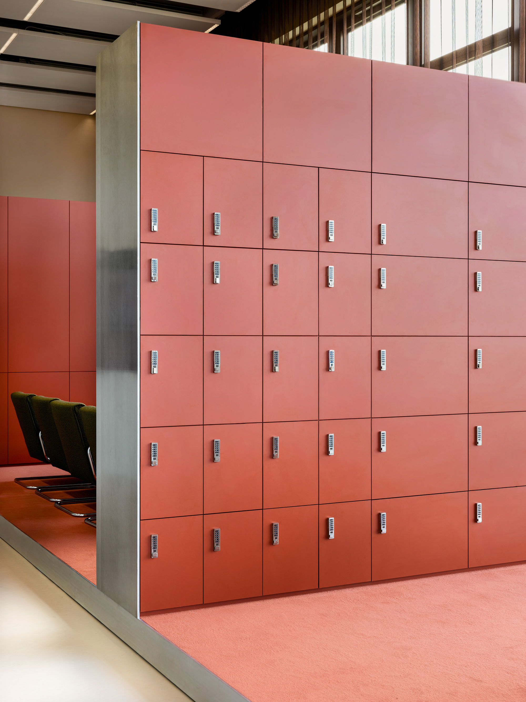 Wall of red lockers with keypad locks, framed by a brushed steel edge, above red carpet and beside green chairs