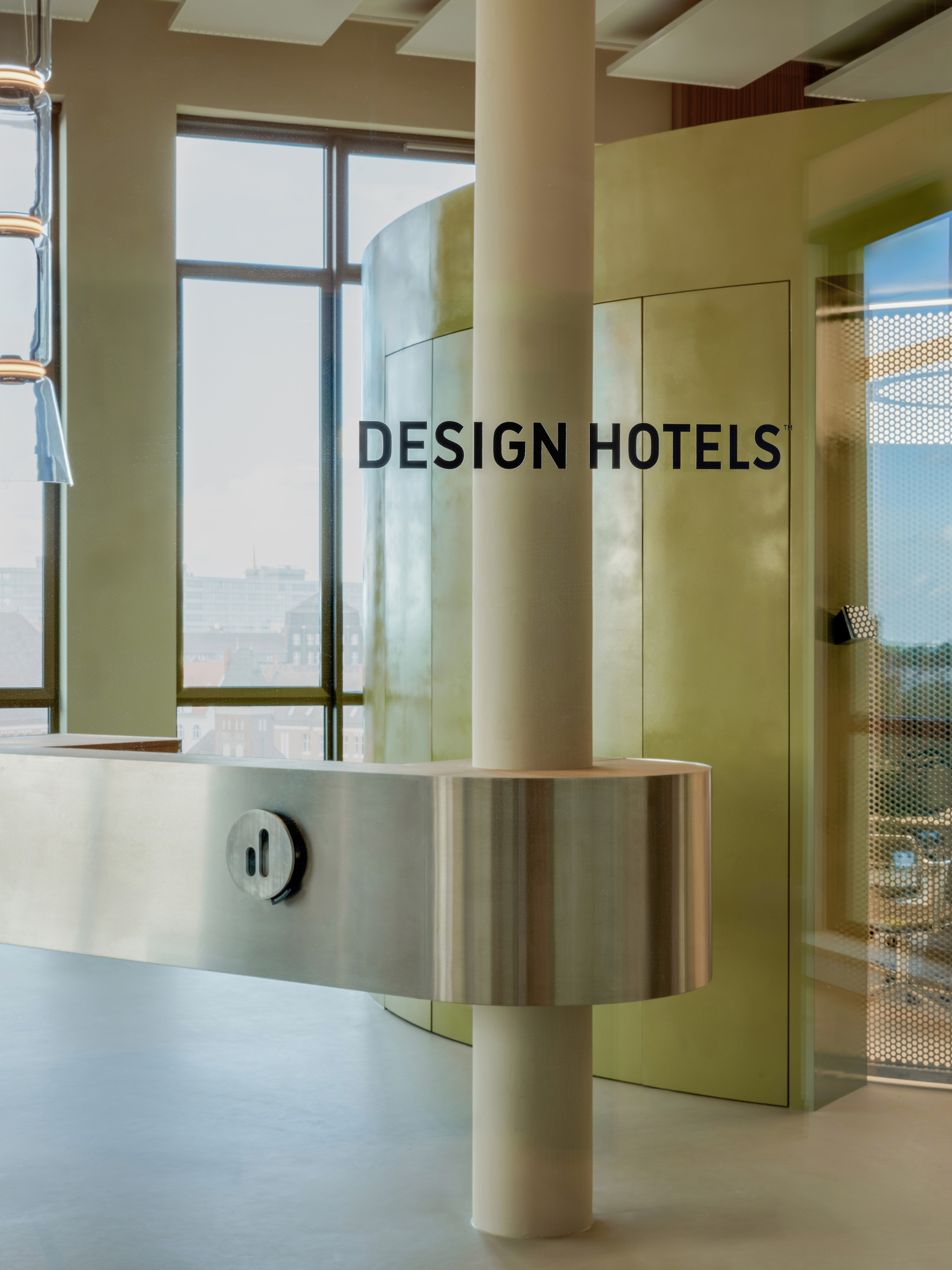 Reception desk in brushed steel wrapping a round column, in front of a curved brass-toned wall with lettering and a window onto the city