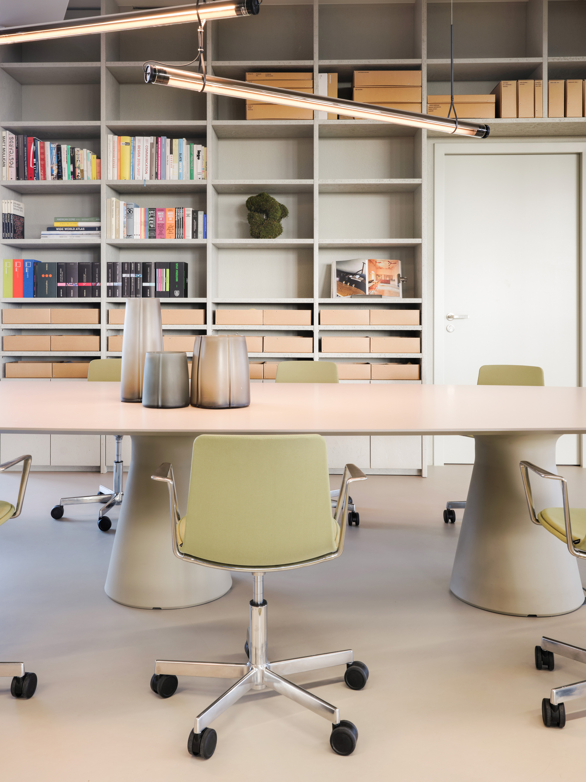 Long meeting table on conical bases with olive swivel chairs, in front of a grey shelving wall of books and boxes