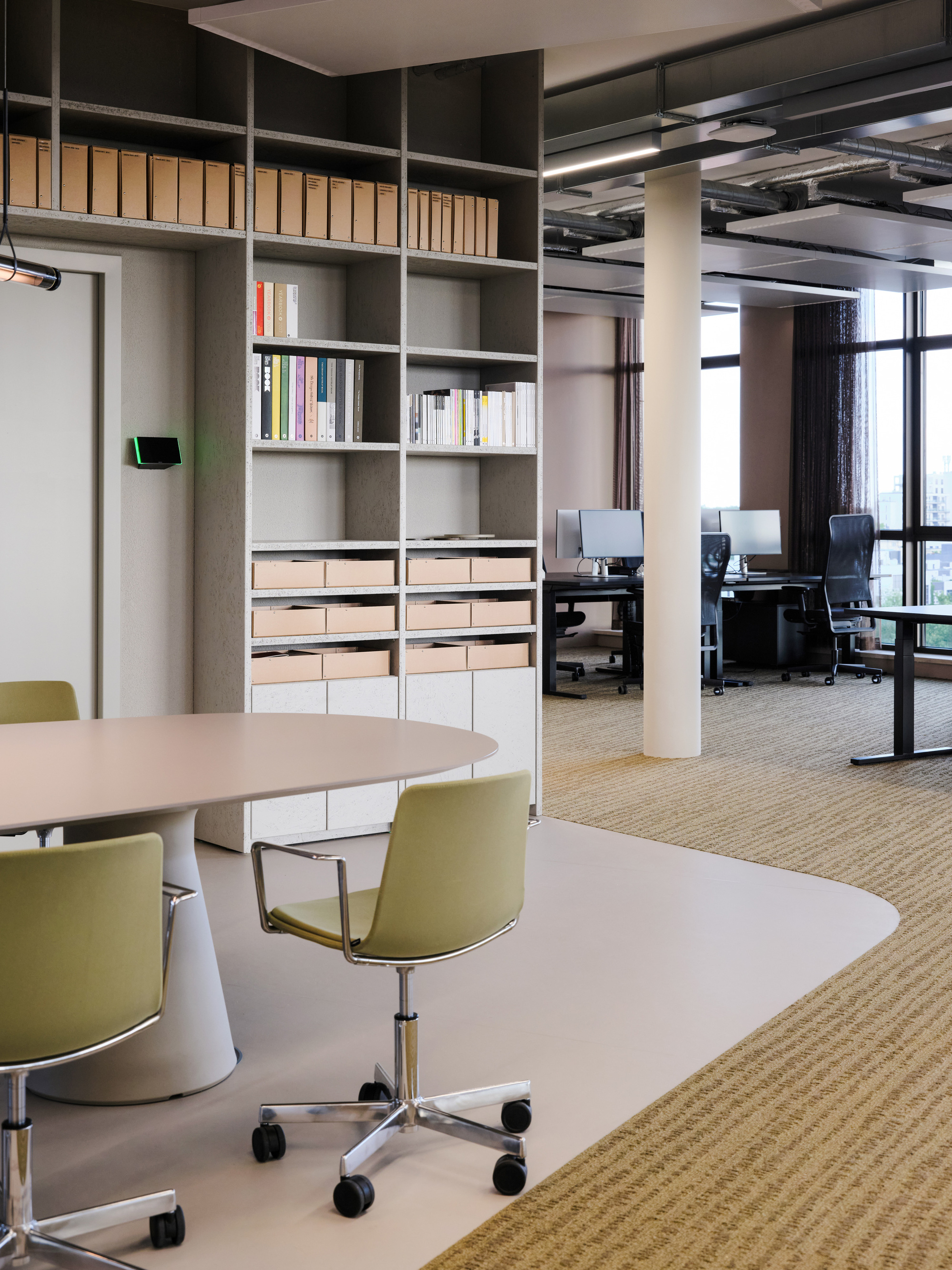 Meeting table and olive chairs on a rounded floor edge, with built-in shelves and a desk area on patterned carpet beyond
