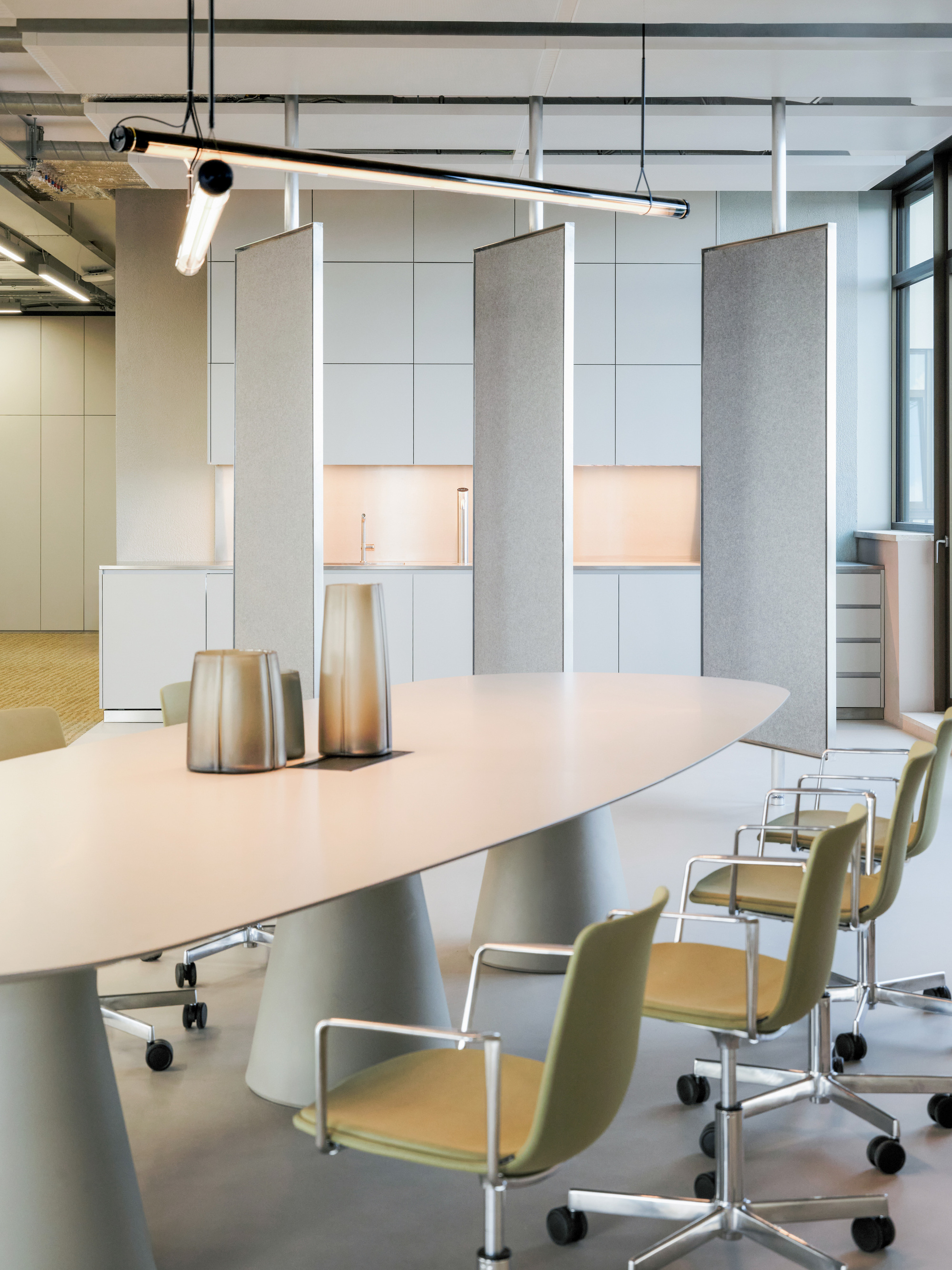 Oval meeting table with olive green chairs and tall grey felt screens in front of a pale kitchen wall