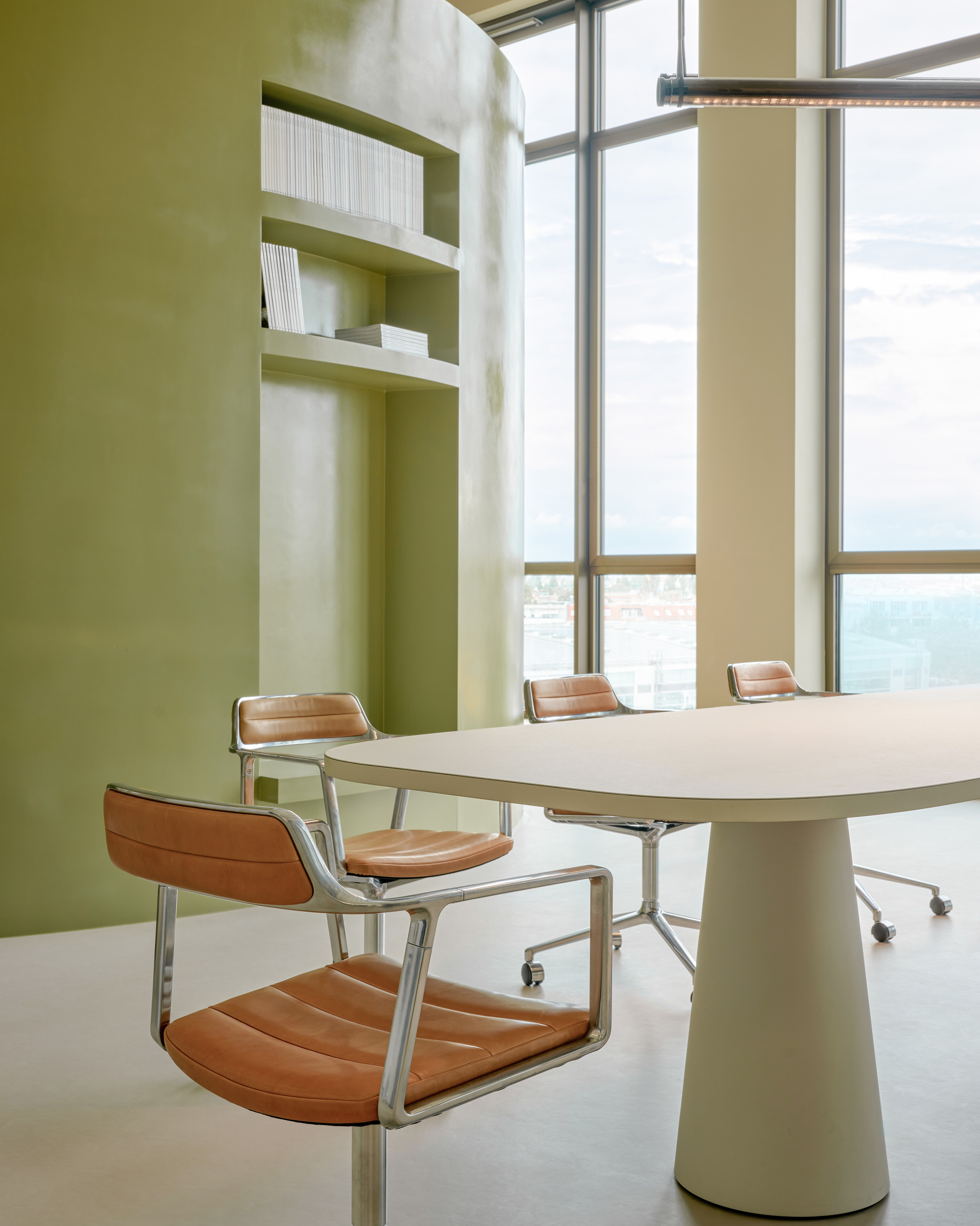Olive green curved wall with built-in bookshelves beside a cream table and tan leather chairs, floor-to-ceiling windows behind under cloudy sky