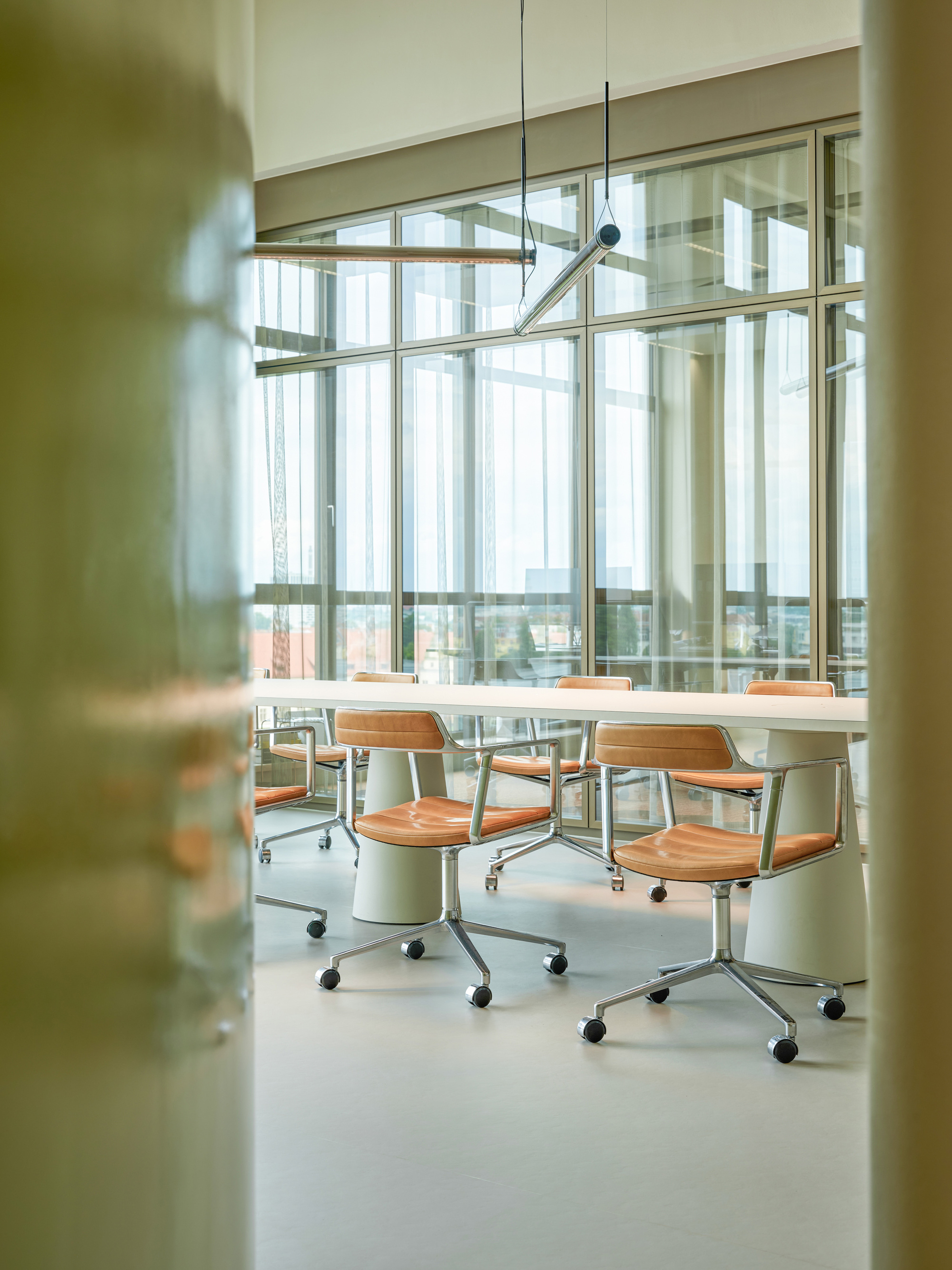 Long table with tan leather chairs seen between two green columns, facing a glazed wall with tall windows in daylight