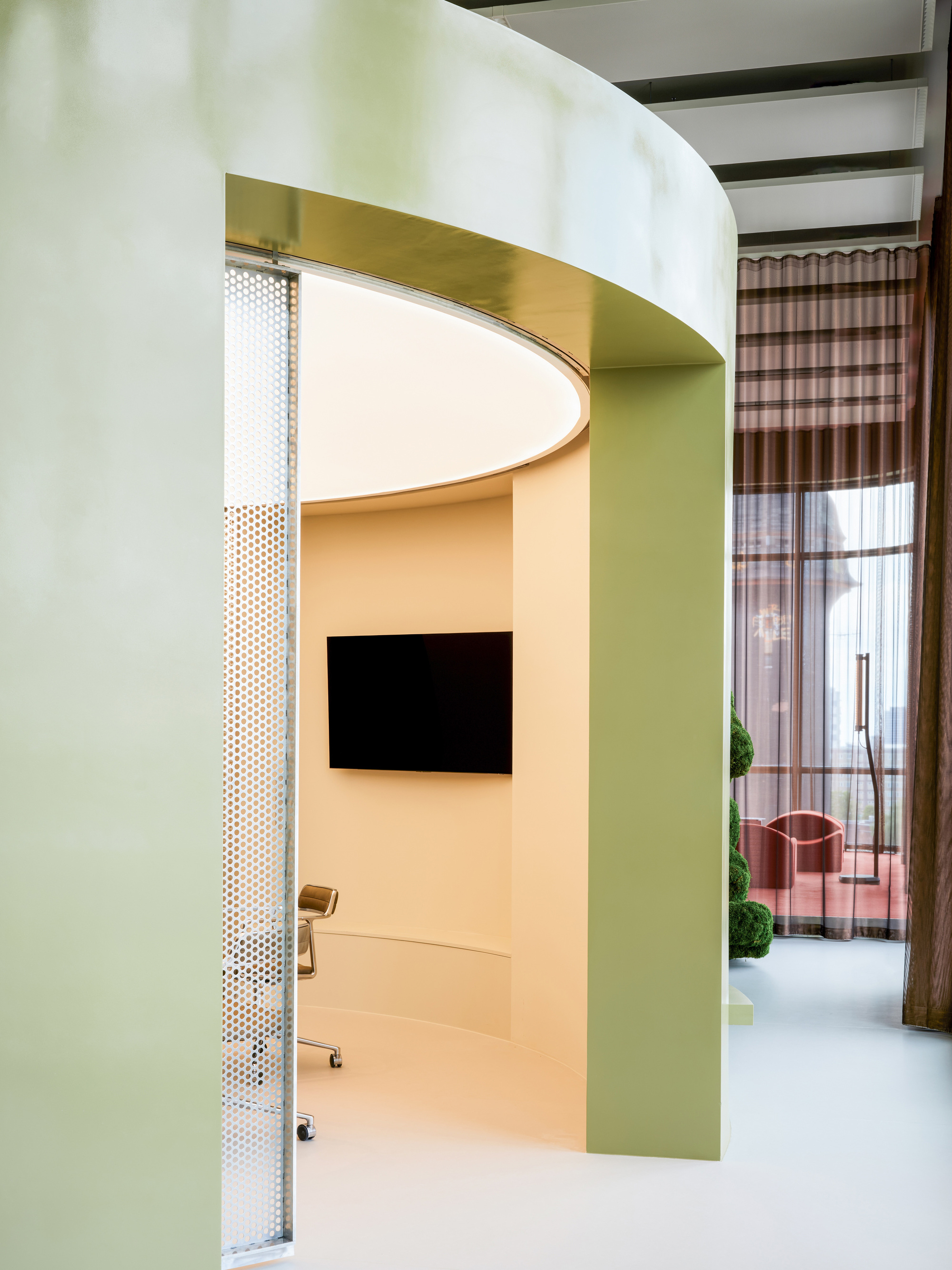 Curved green meeting pod with a round ceiling light and wall-mounted screen, a sheer brown curtain and potted topiary beside it