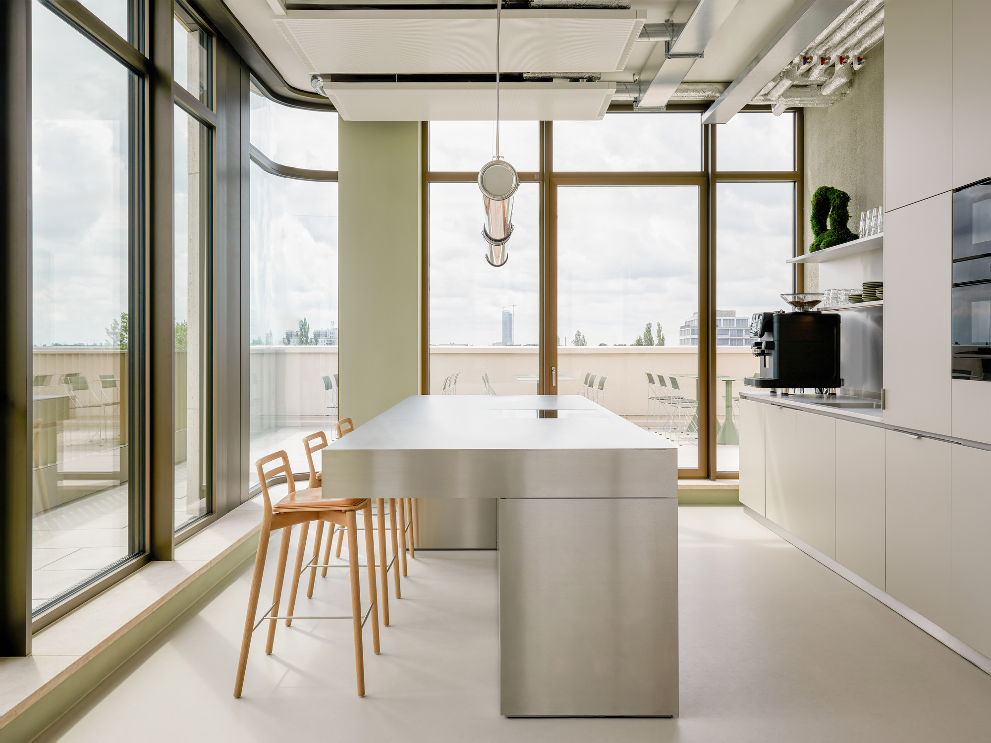 Stainless steel kitchen island with timber stools and a coffee machine, facing a glazed corner and roof terrace under overcast sky