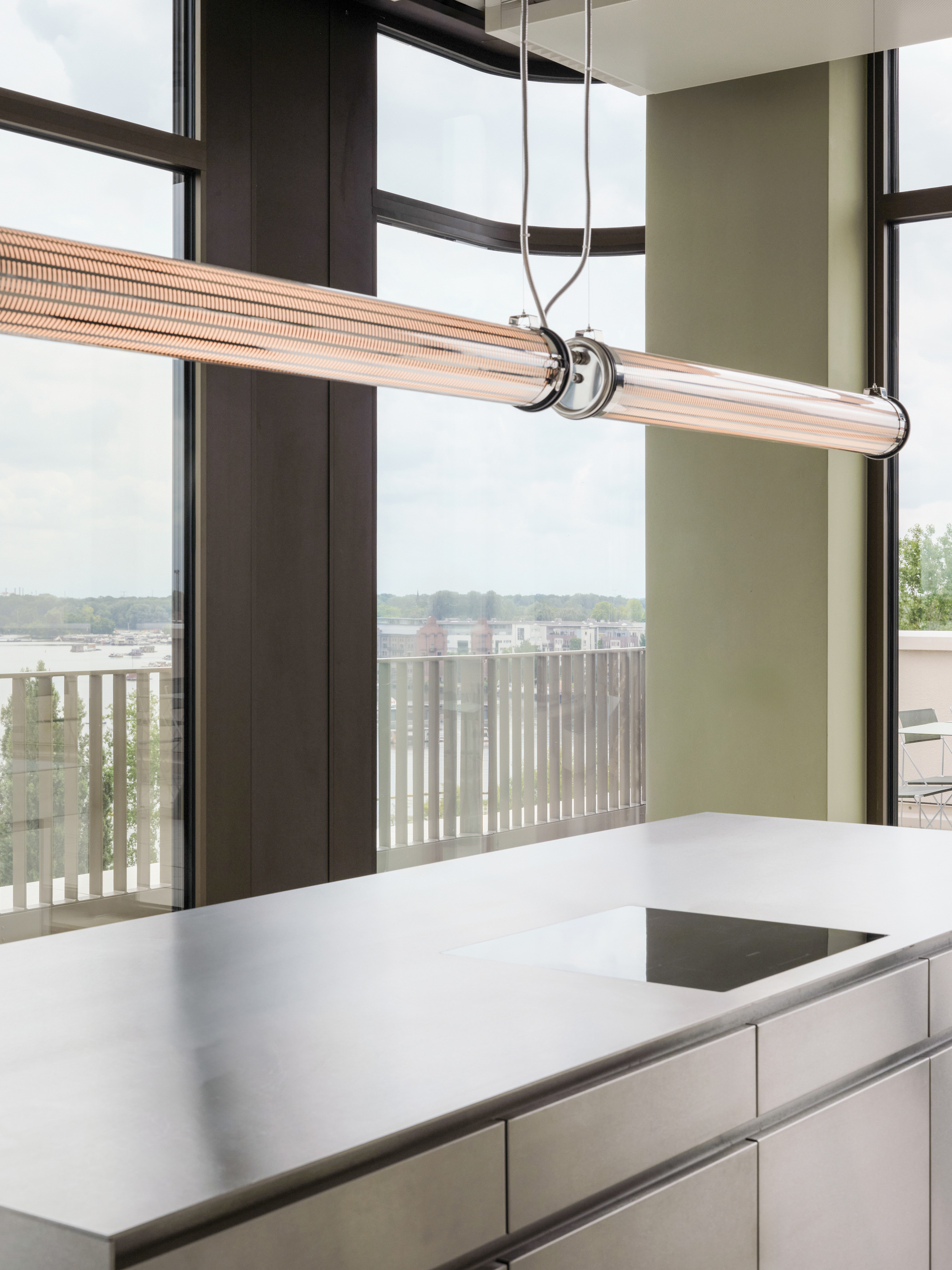 Suspended glass tube pendant light above a stainless steel kitchen island, with tall windows and a slatted balcony railing under an overcast sky
