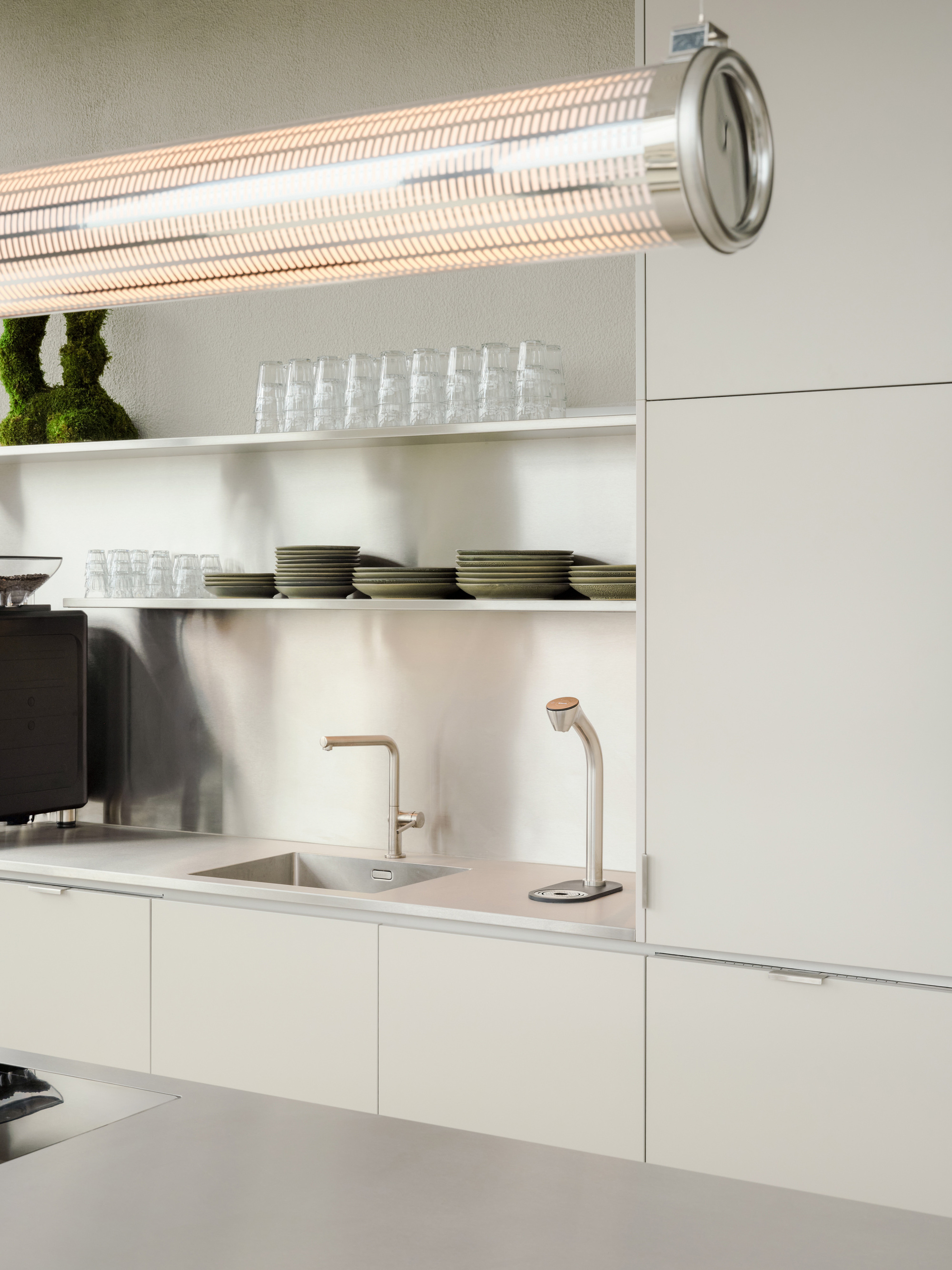 Stainless steel kitchen wall with open shelves of glasses and plates, a sink, a tap and a tube pendant glowing overhead