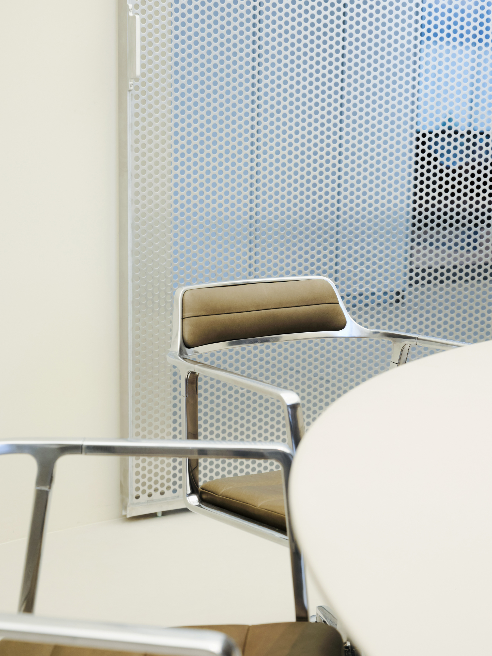 Chrome-framed armchair with taupe leather upholstery beside a round white table, set against a perforated metal screen with blue light behind
