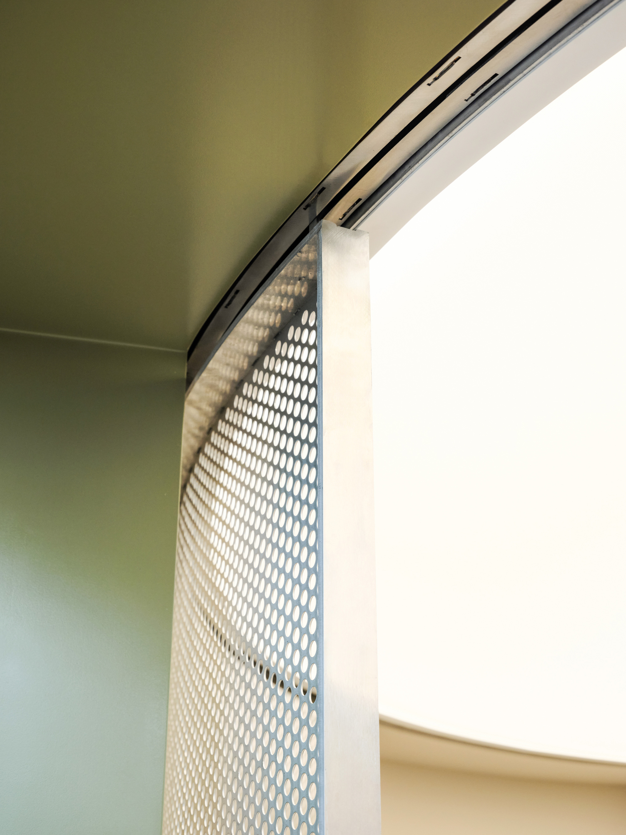 Curved perforated metal panel hanging from a ceiling track beside a bright window, with olive green walls in soft light