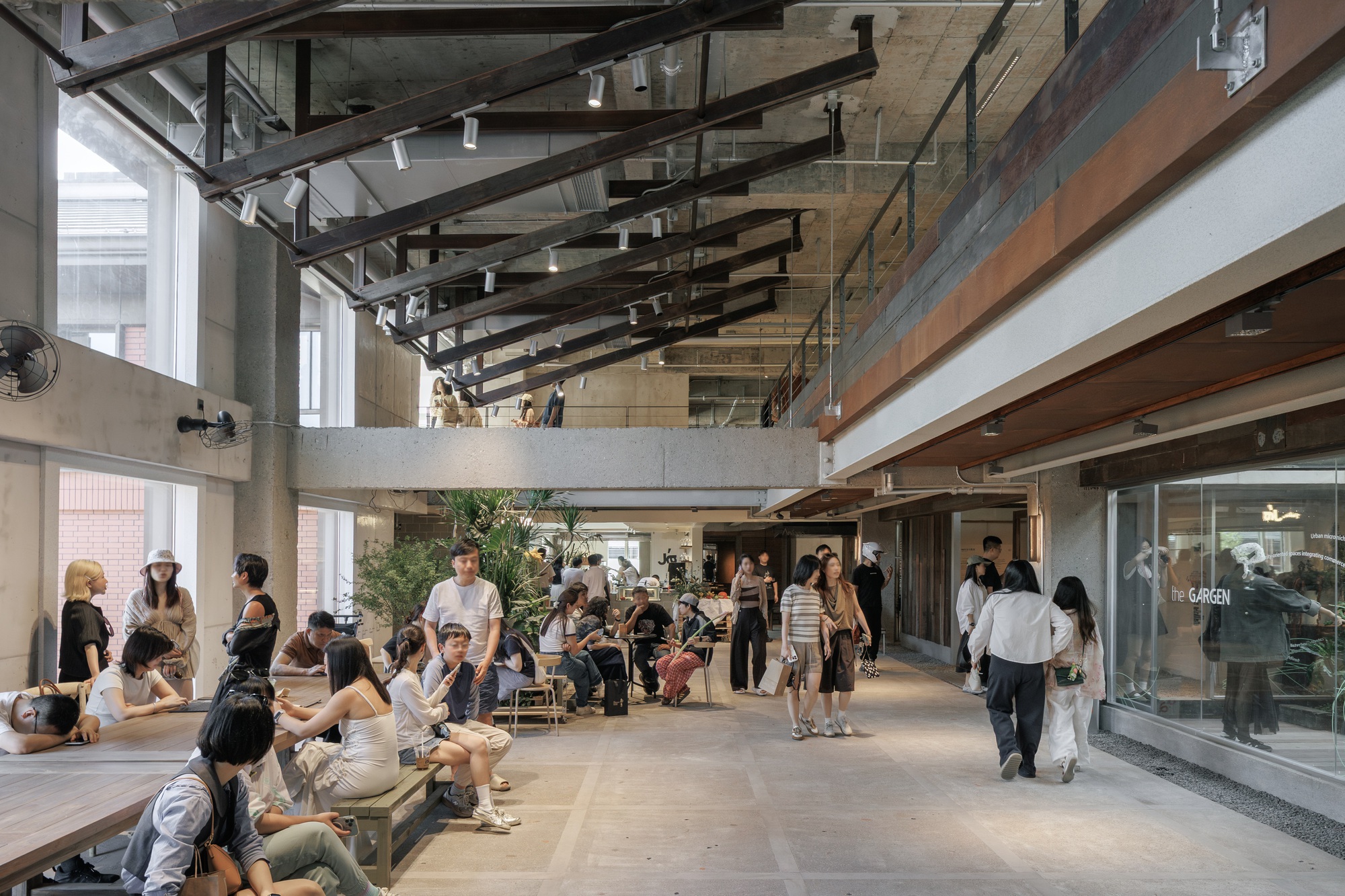 Crowded ground-floor concourse with diagonal steel rafters, concrete beams and visitors seated on timber benches beneath a mezzanine