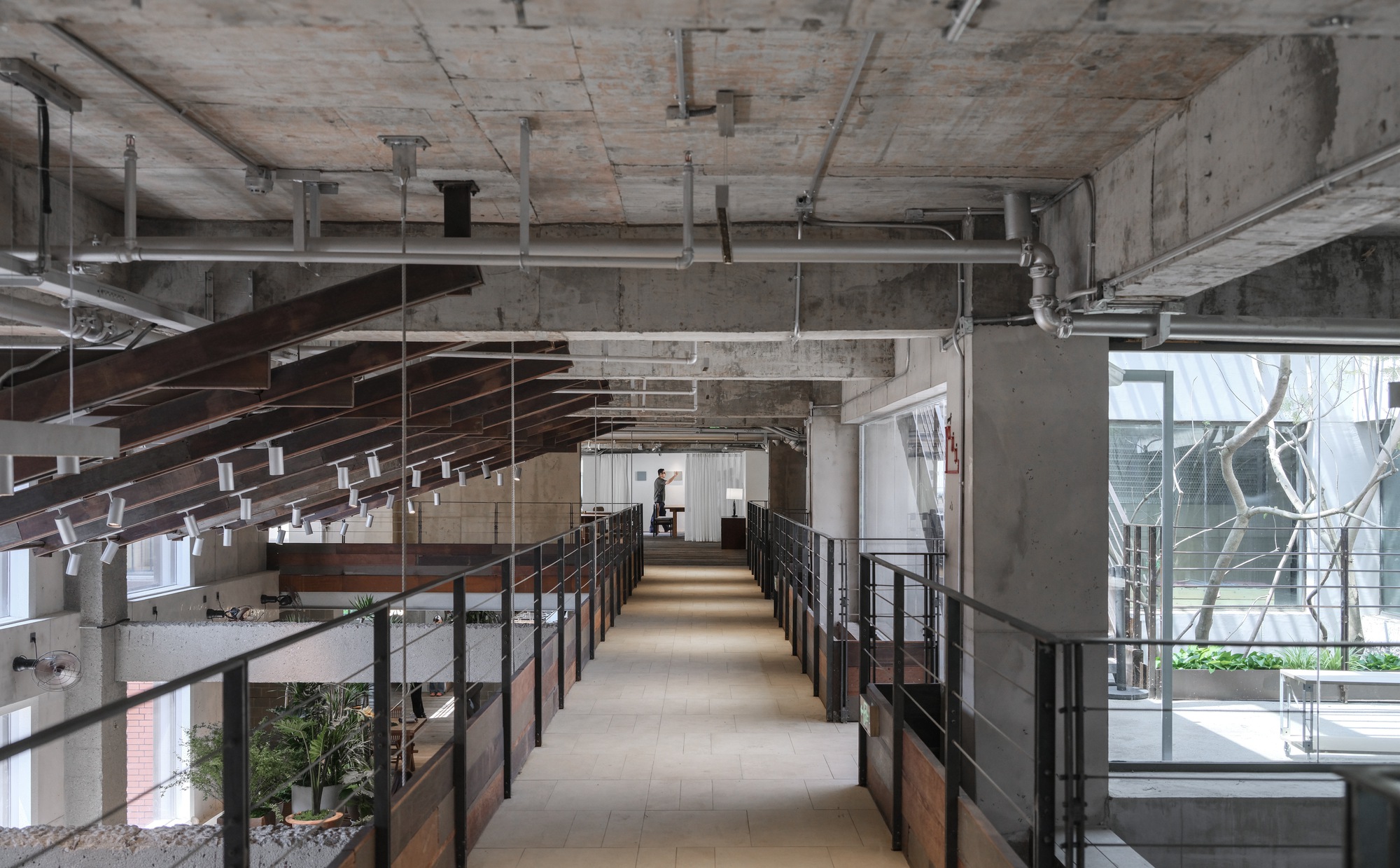 Upper-level walkway lined with steel railings and timber-clad parapets, with track lights hanging from rusted rafters toward a bright window