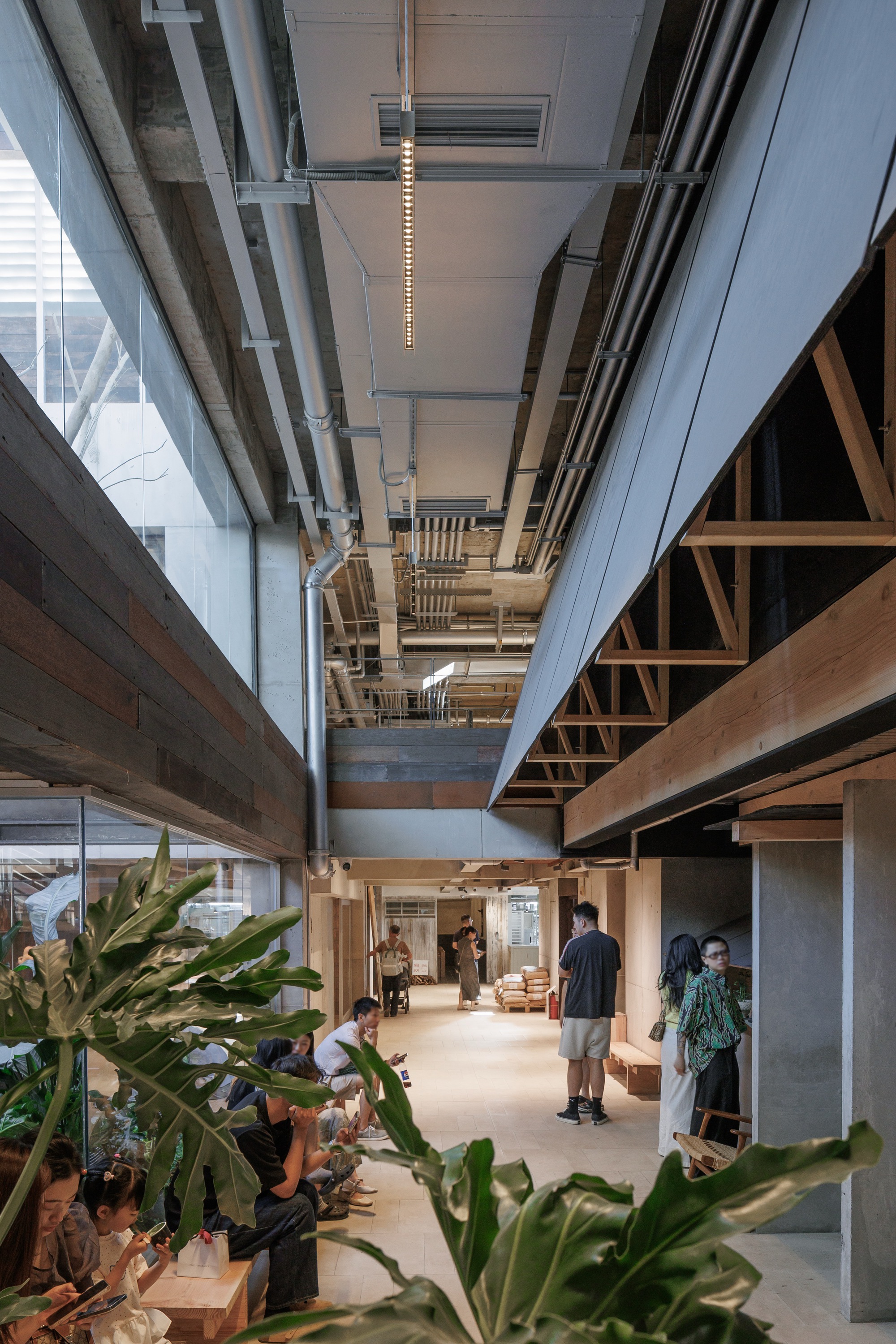 Narrow interior passage under exposed ducts, with monstera leaves in the foreground and visitors sitting along a timber-clad wall