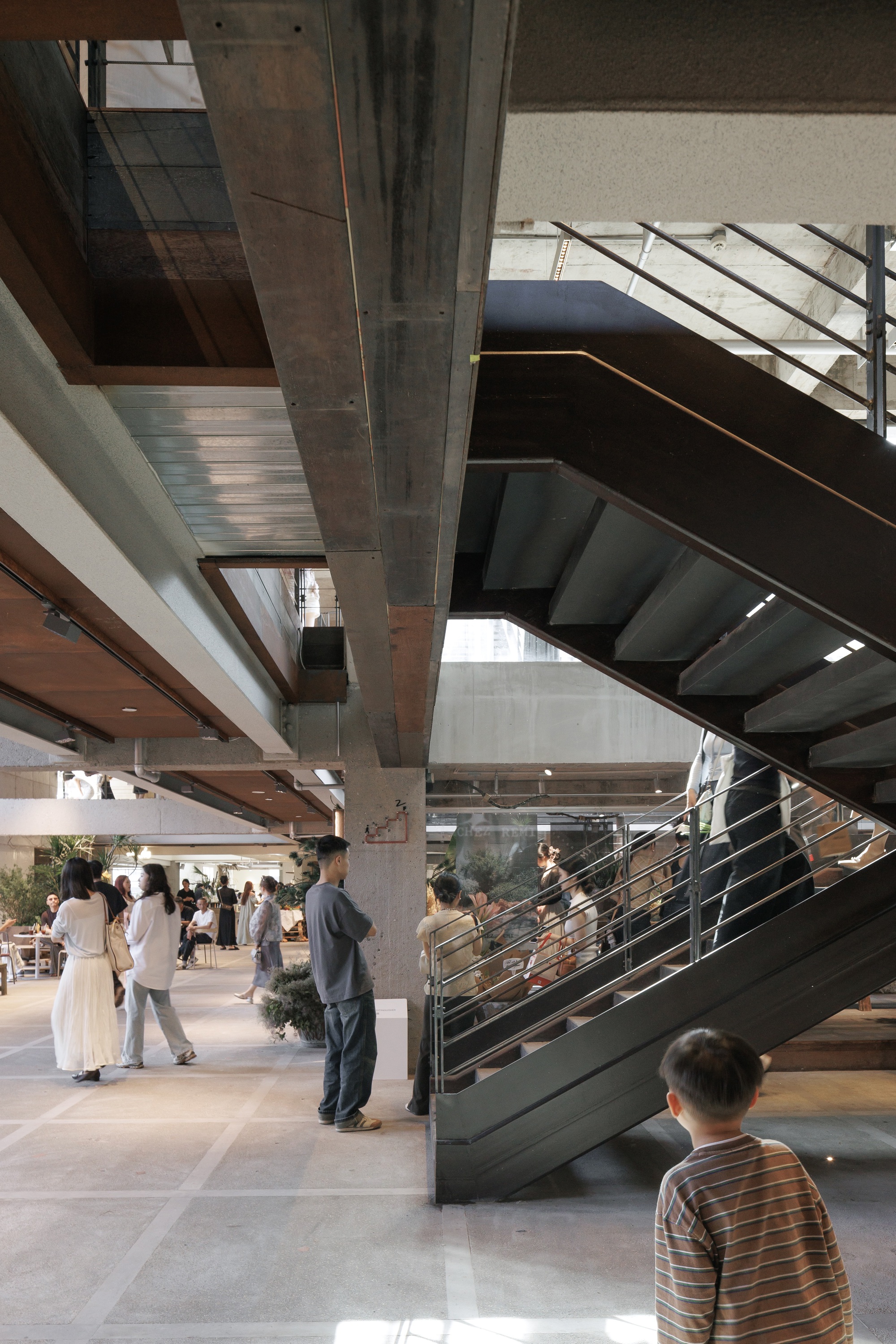 Dark steel staircase beside a concrete column, with a boy in a striped shirt watching visitors cross the floor