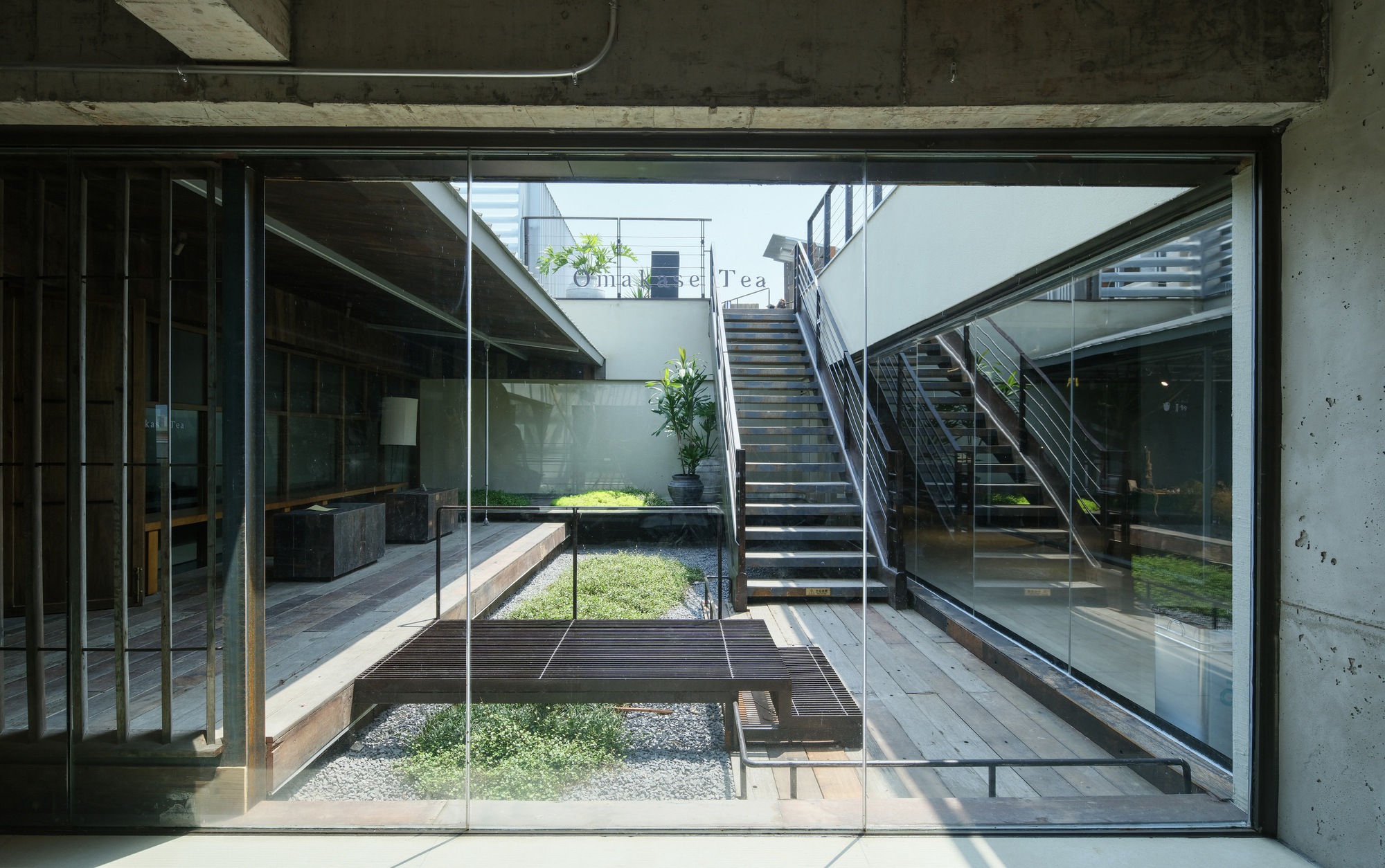 Glazed opening onto a sunlit courtyard with gravel, a steel bench, groundcover plants and a stair rising to an upper terrace
