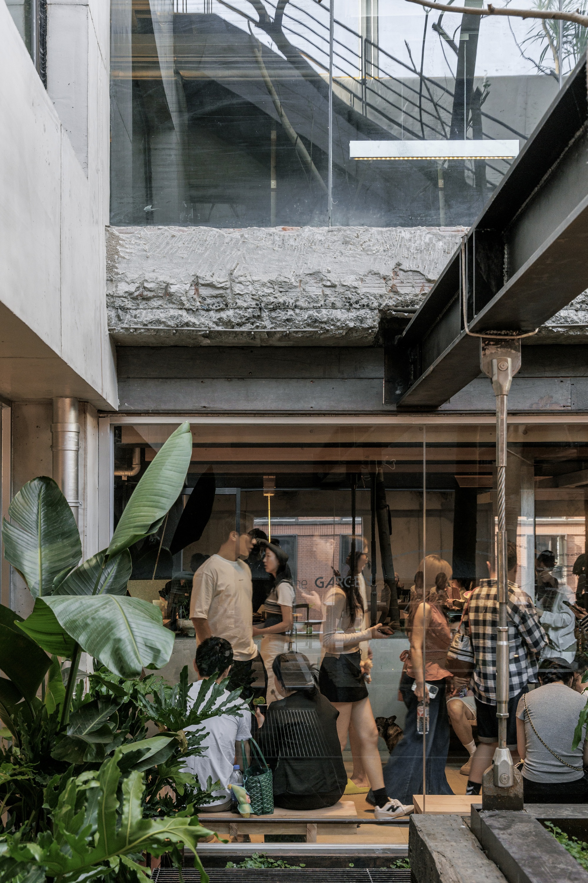 Glazed courtyard edge where visitors sit on a wooden bench behind banana leaves, beneath a steel beam and rough concrete slab