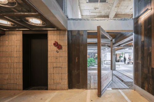 Pivoting timber door opening from a brick-tiled elevator lobby onto a plank-floored courtyard with people in the distance