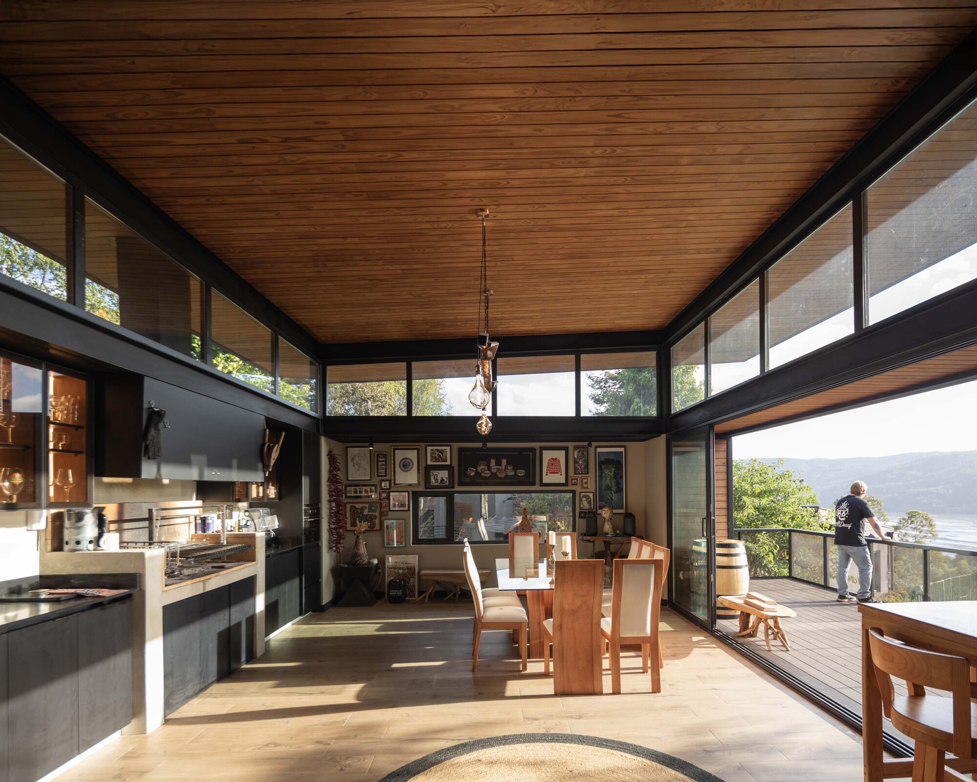Open kitchen and dining room under a timber plank ceiling, with a man on the deck beyond the sliding glass doors in afternoon light
