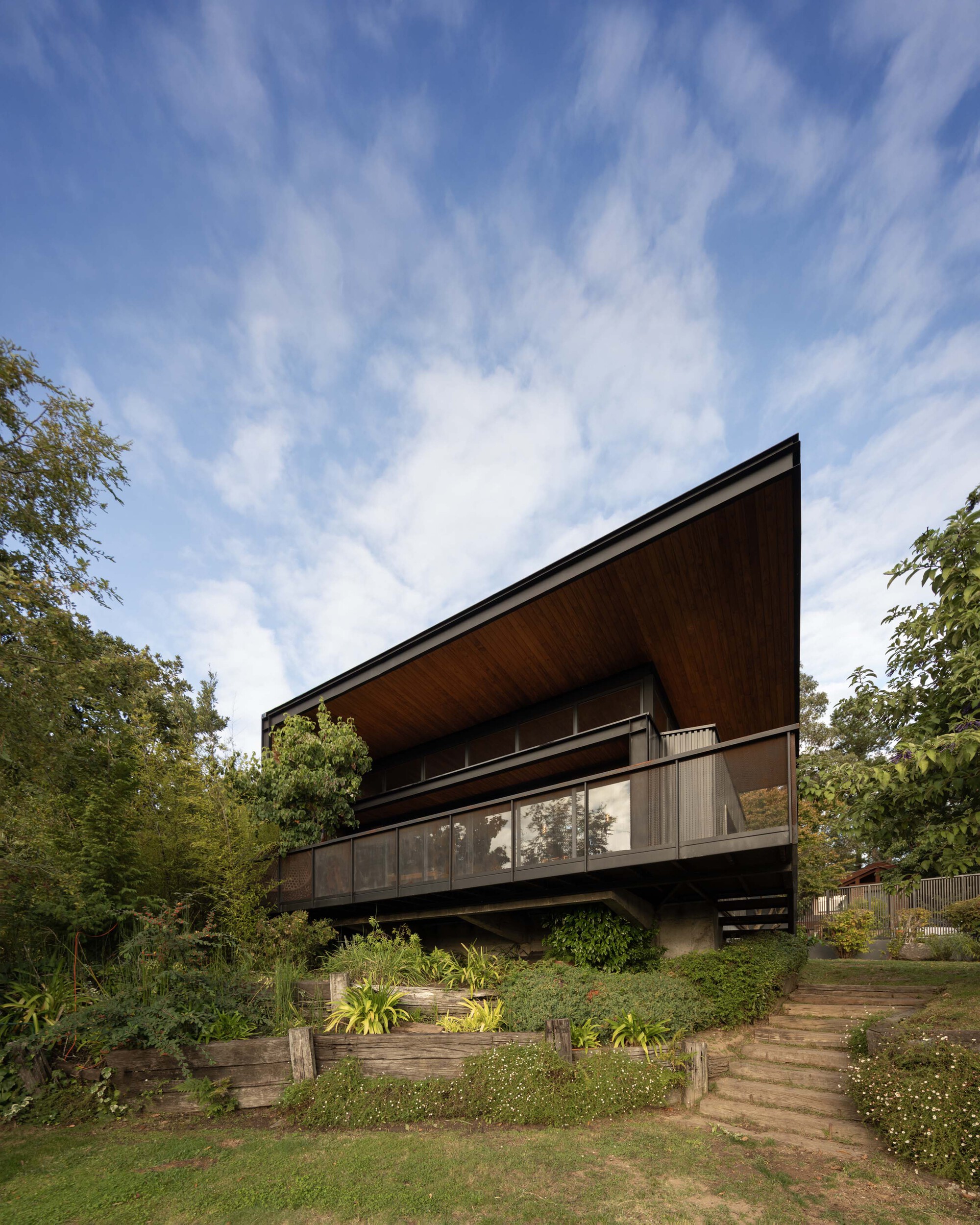 Low-angle view of the sloping timber-lined roof and glazed balcony above a planted terraced slope under wispy clouds