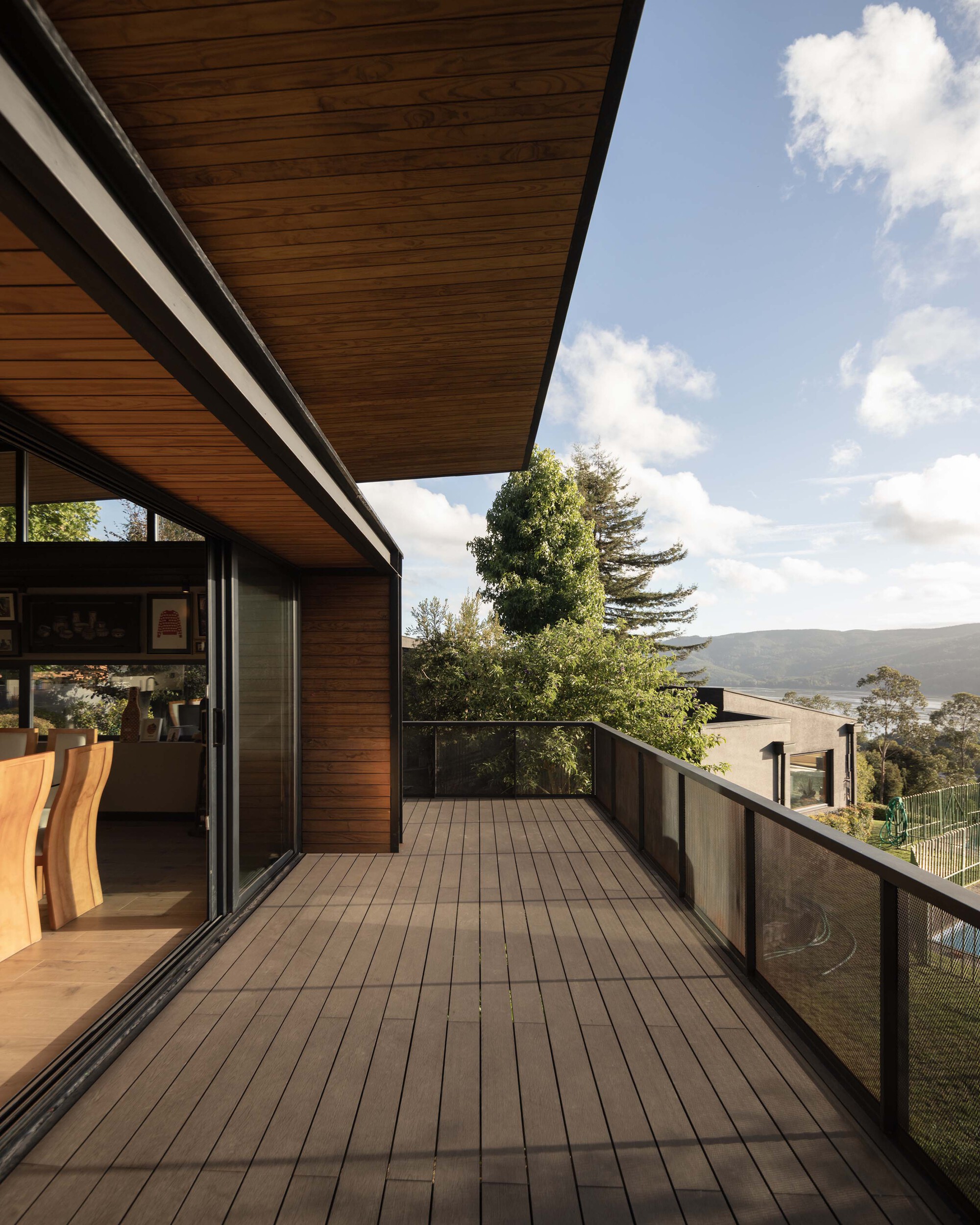 Timber deck running along the sliding glass wall beneath a deep roof overhang, with tall trees and distant hills under scattered clouds