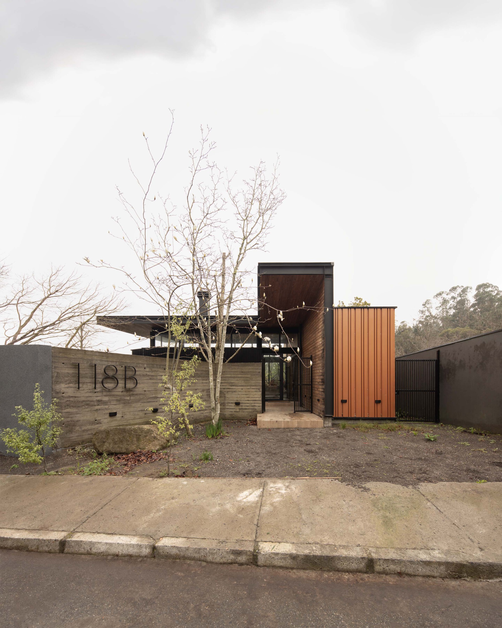 Street facade with a board-formed concrete wall, house number, orange metal-clad volume and a bare tree under an overcast sky