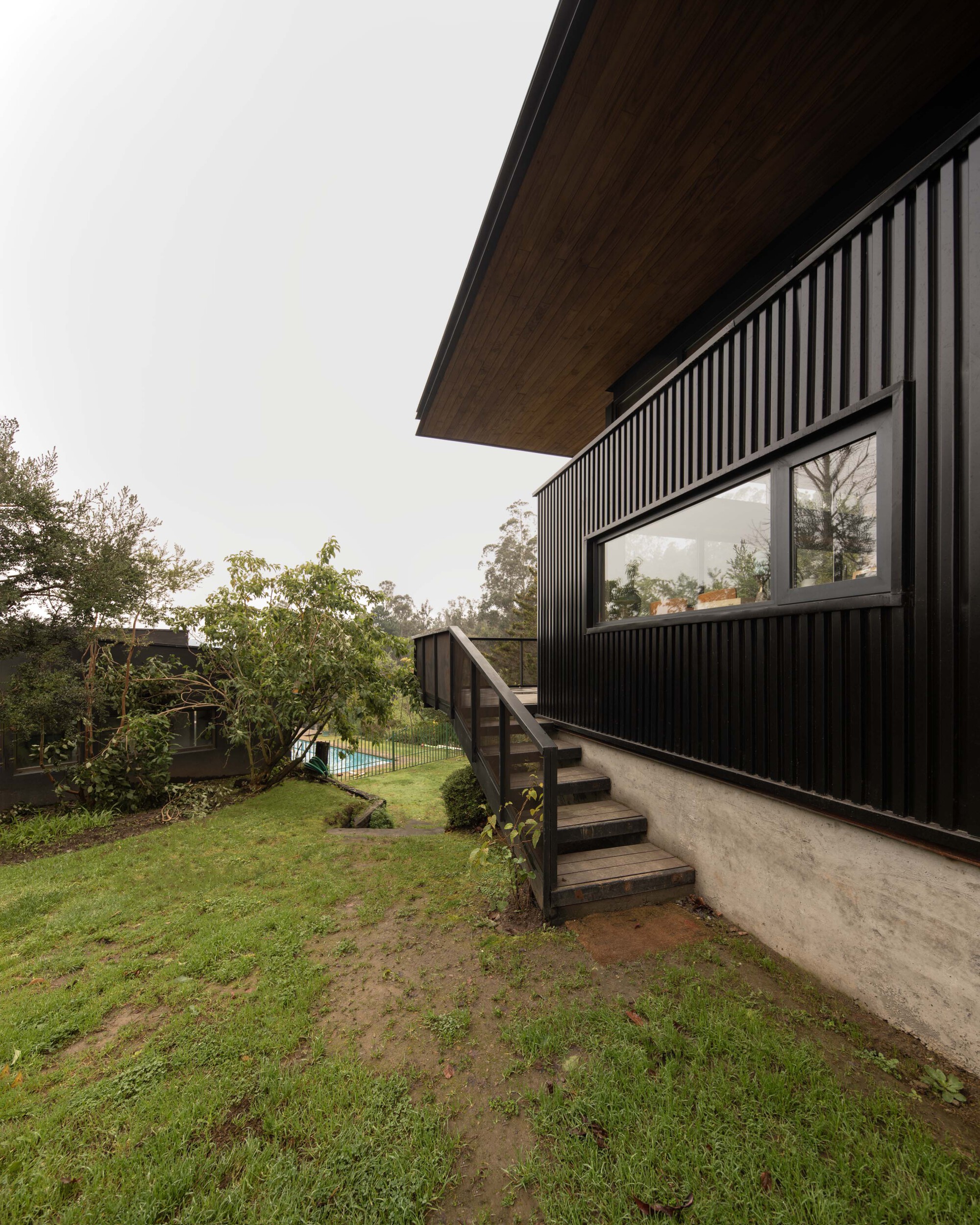 Black vertical-clad side wall with a ribbon window and timber steps leading down to a wet lawn under grey sky