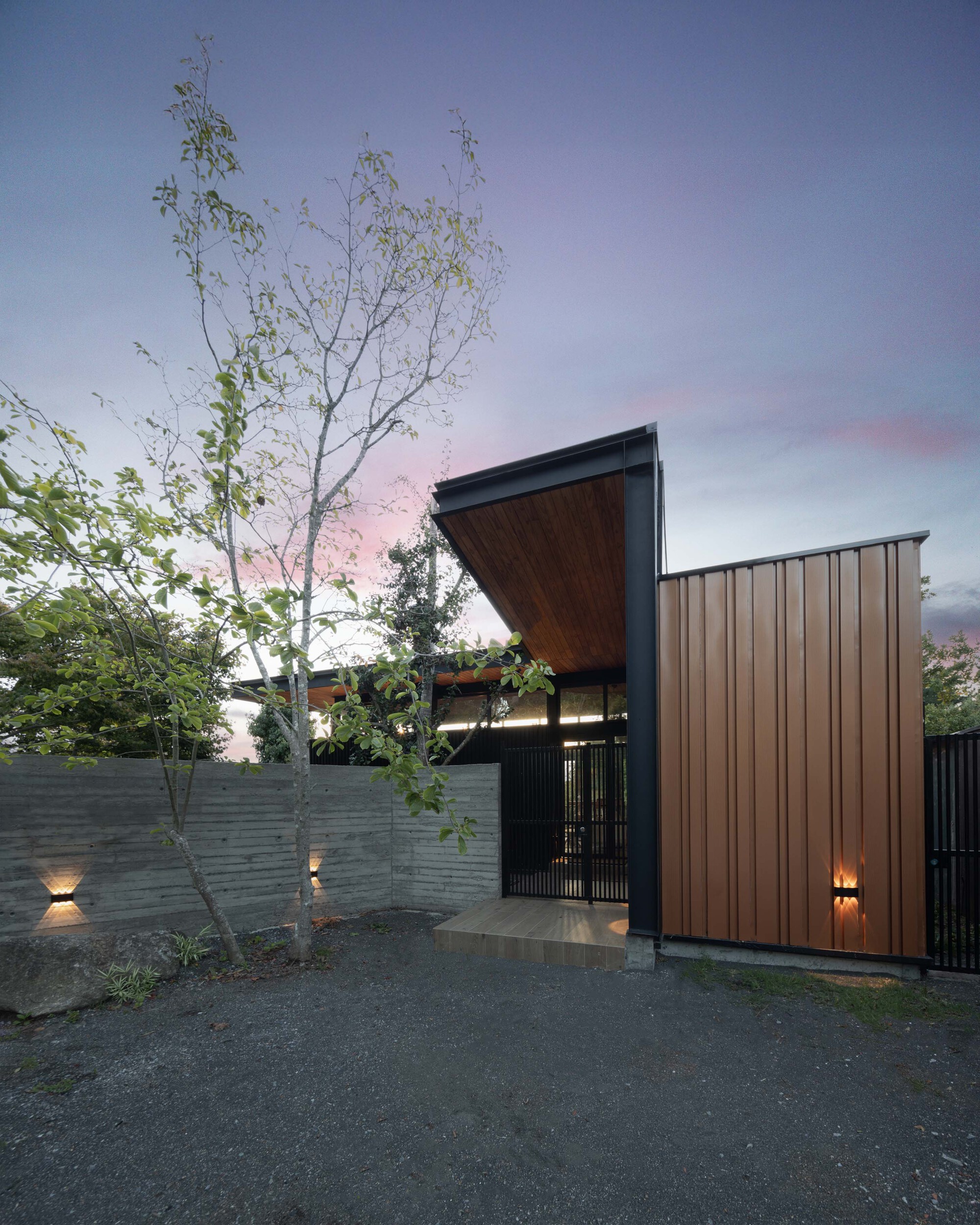 Entry gate beneath a cantilevered timber-lined roof, beside a board-formed concrete wall and copper-toned vertical cladding, with wall lights glowing at pink du