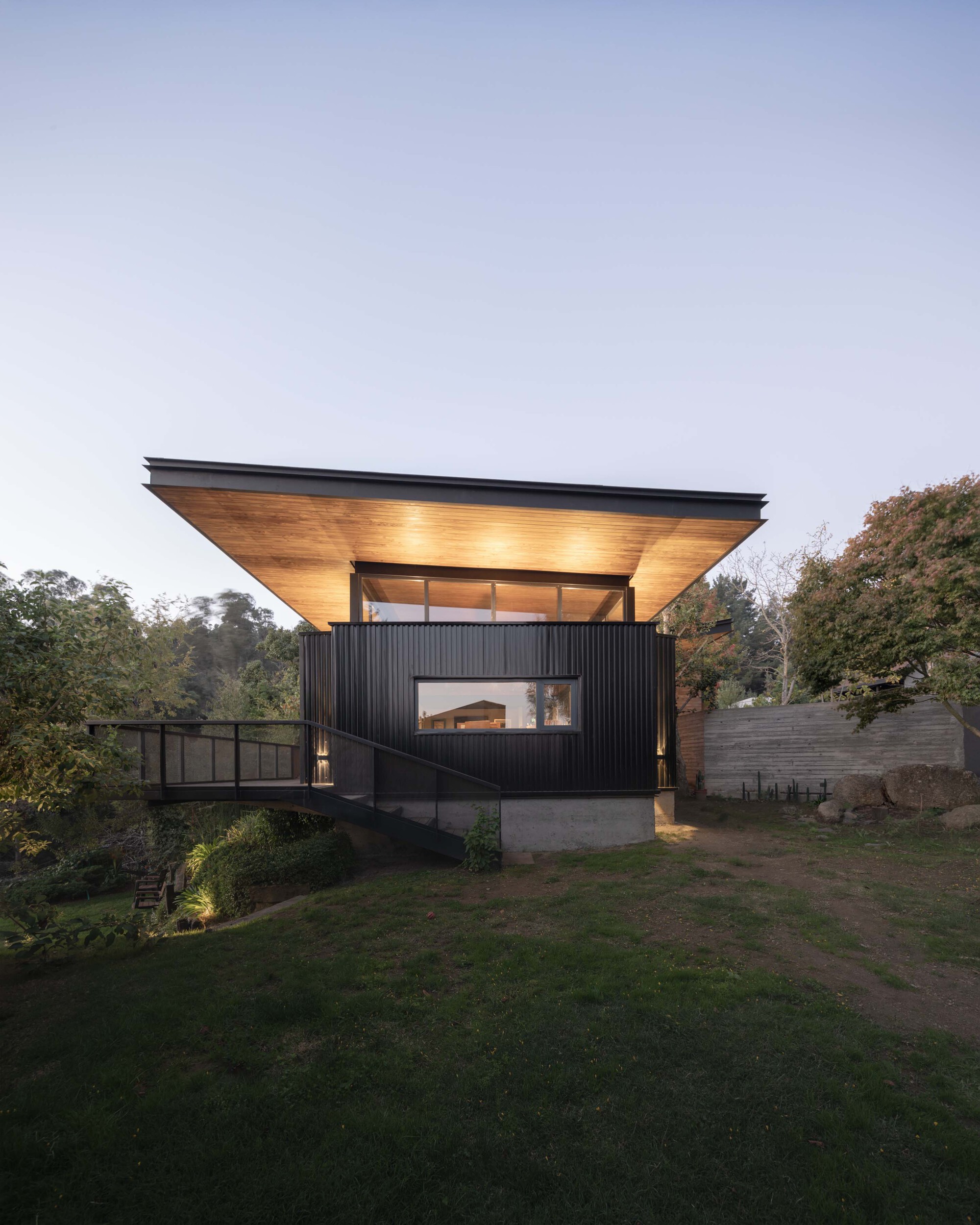 Black corrugated volume under a wide floating timber-soffit roof, reached by a steel footbridge across a grassy slope at dusk