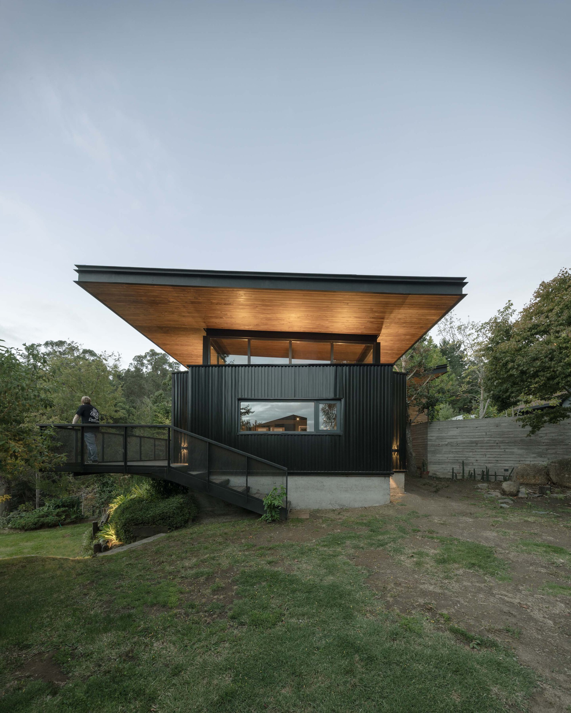 Side view of the dark clad pavilion and glass-railed footbridge, with a person standing at the railing under an overcast evening sky