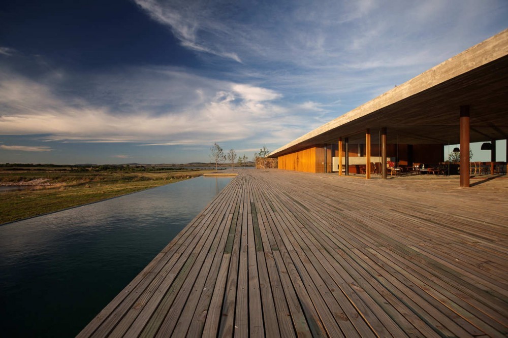 Long plank deck running beside a still pool toward a covered porch with slender timber columns under a cloudy sky