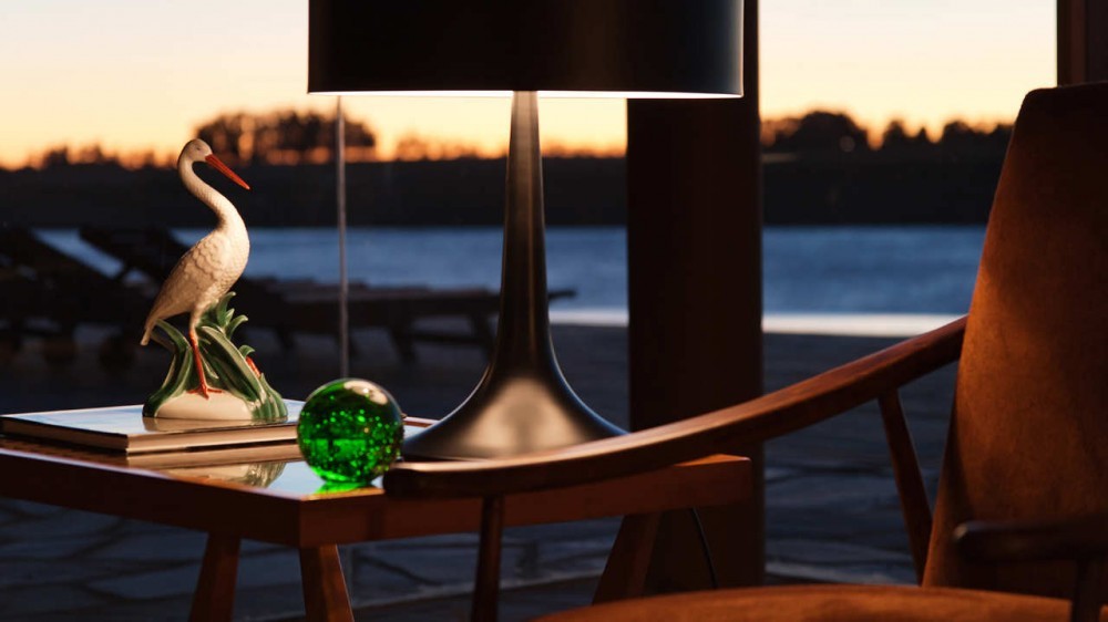 Close-up of a black table lamp, porcelain stork figurine and green glass ball beside a wooden armchair at dusk