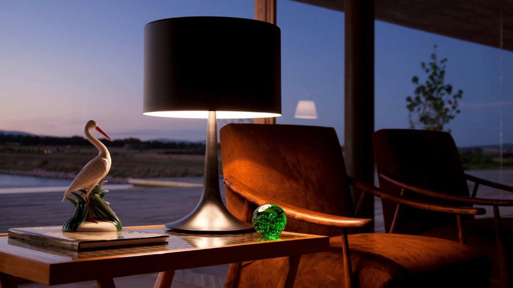Table lamp and stork figurine on a side table next to rust-colored armchairs facing a glass wall at twilight
