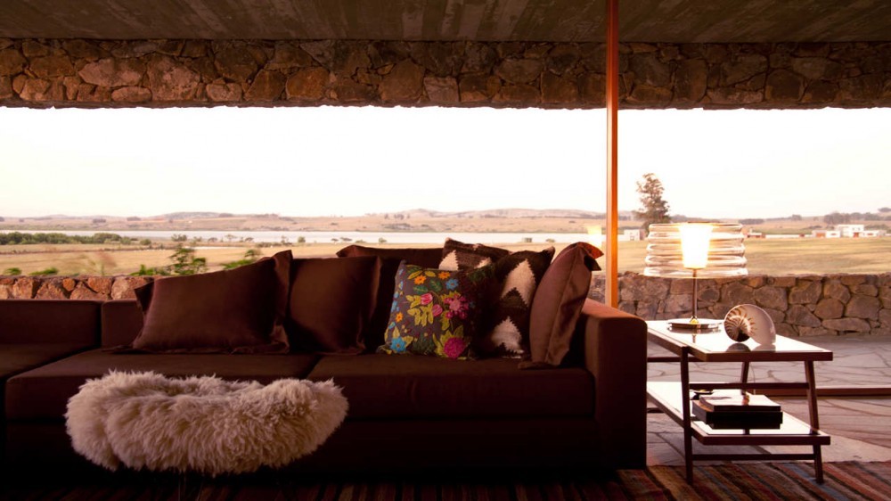 Brown sofa with patterned cushions and a sheepskin beneath a stone-faced lintel, opening to a wide pastoral landscape at dusk