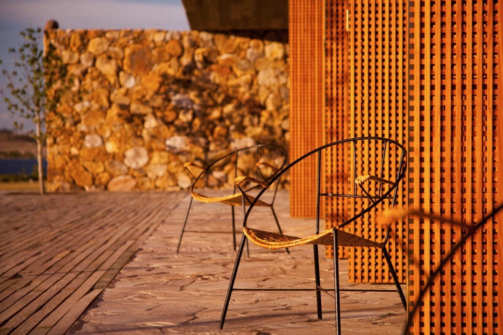 Black steel and woven-seat chairs on a stone terrace, with a rubble stone wall and timber lattice screens in low golden light