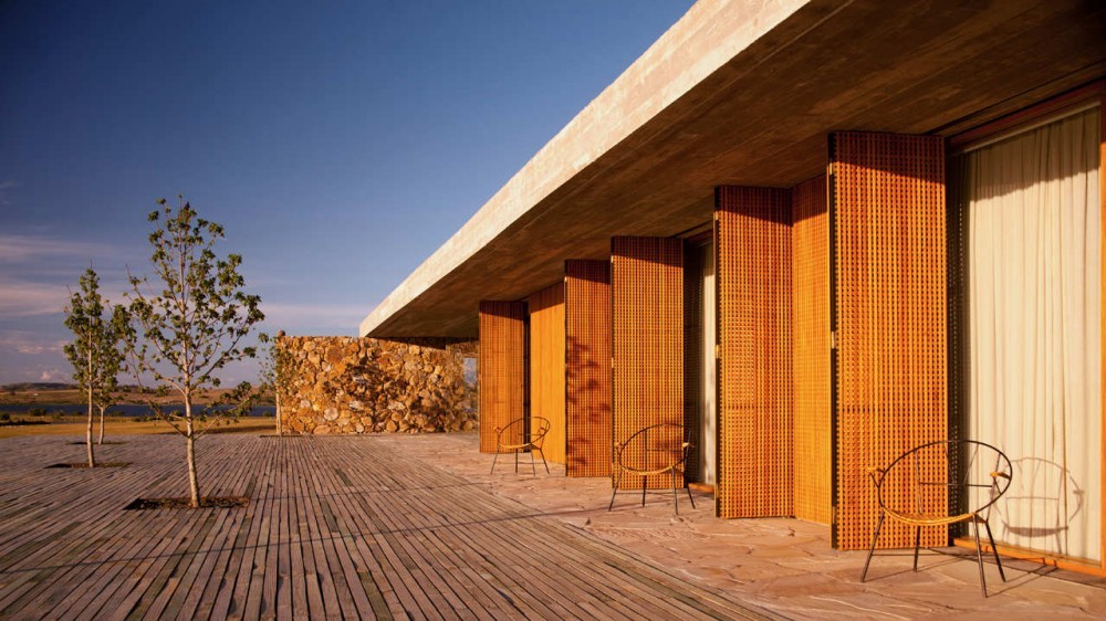 Row of folding timber lattice shutters under a concrete roof slab, with chairs on a deck and young trees at sunset